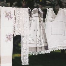 Assorted white linens, lace, and embroidered textiles hang on a line outdoors against a blurred green background.