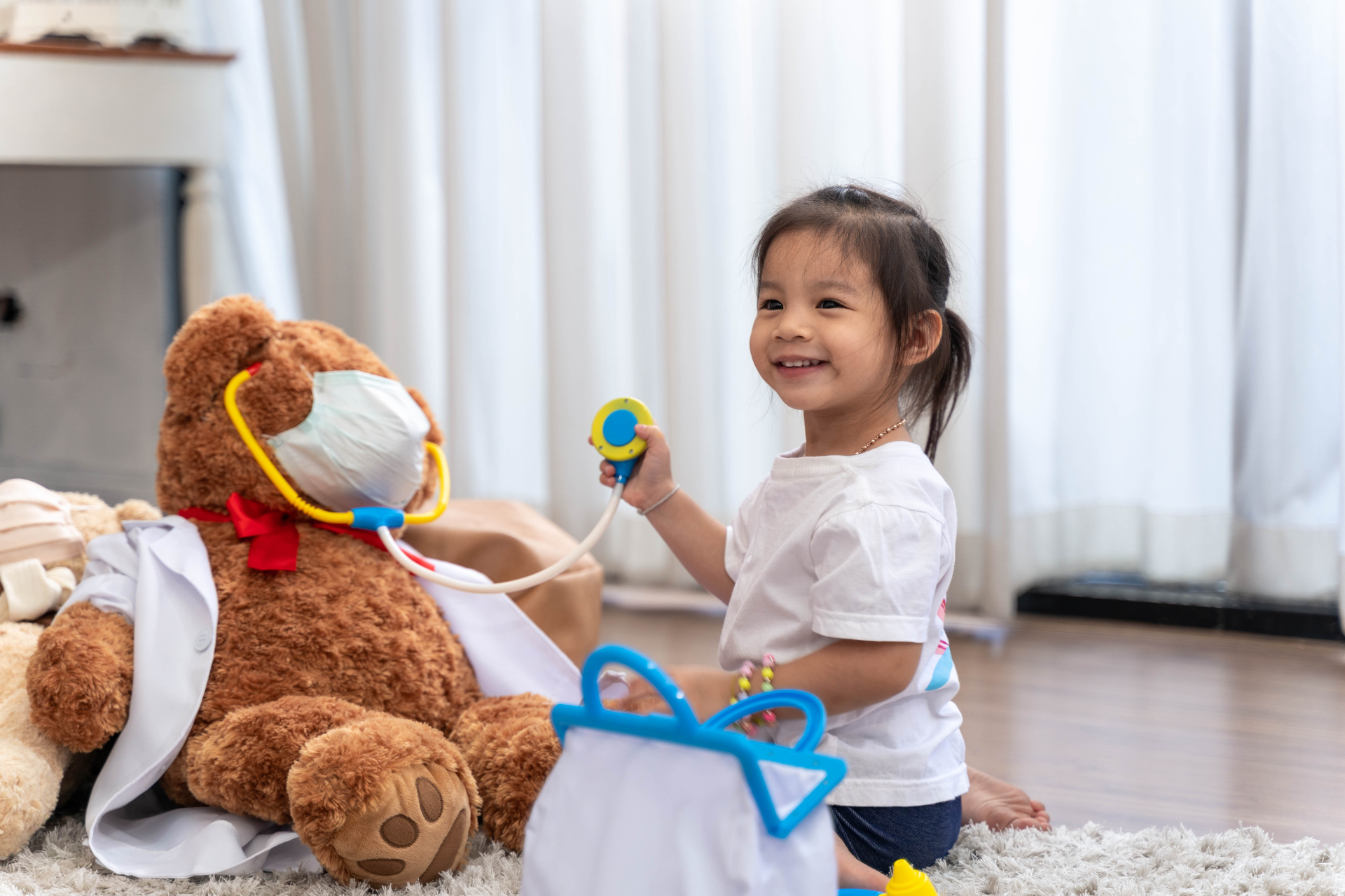 Mädchen spielt Arzt mit einem Teddybären, der ein Stethoskop hält. Sie lächelt. Drinnen, weisse Vorhänge.