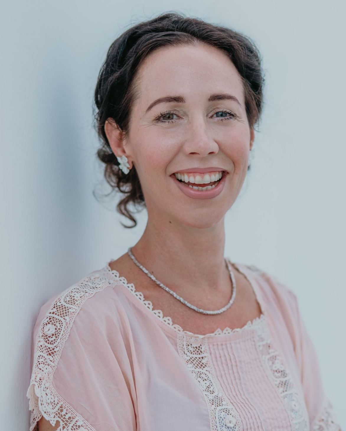 Lächelnde Frau mit dunklem Haar, rosa Oberteil und Halskette, vor einer weißen Wand.