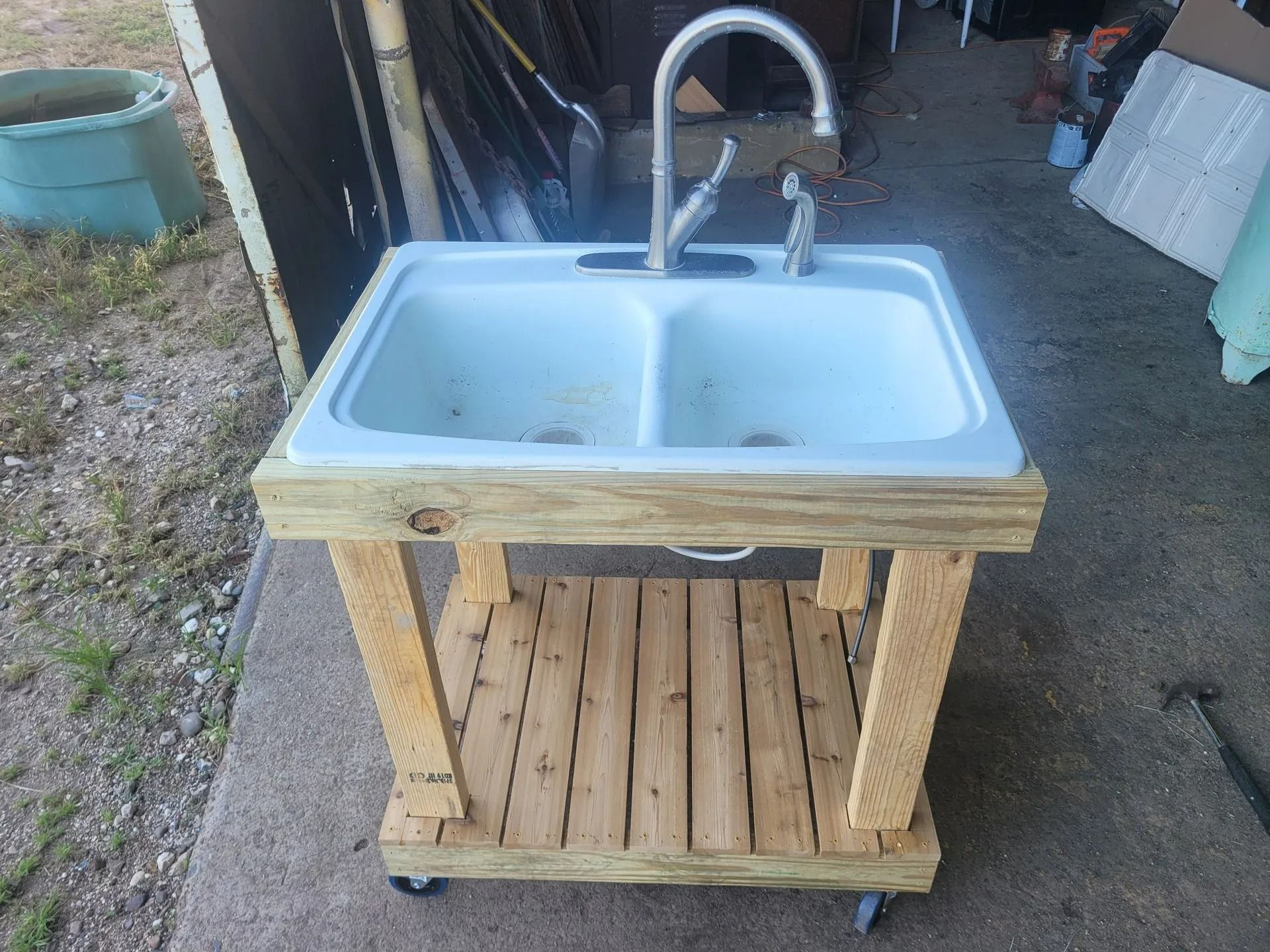 Outdoor sink with double basins, mounted on a wooden frame with a lower shelf and casters.