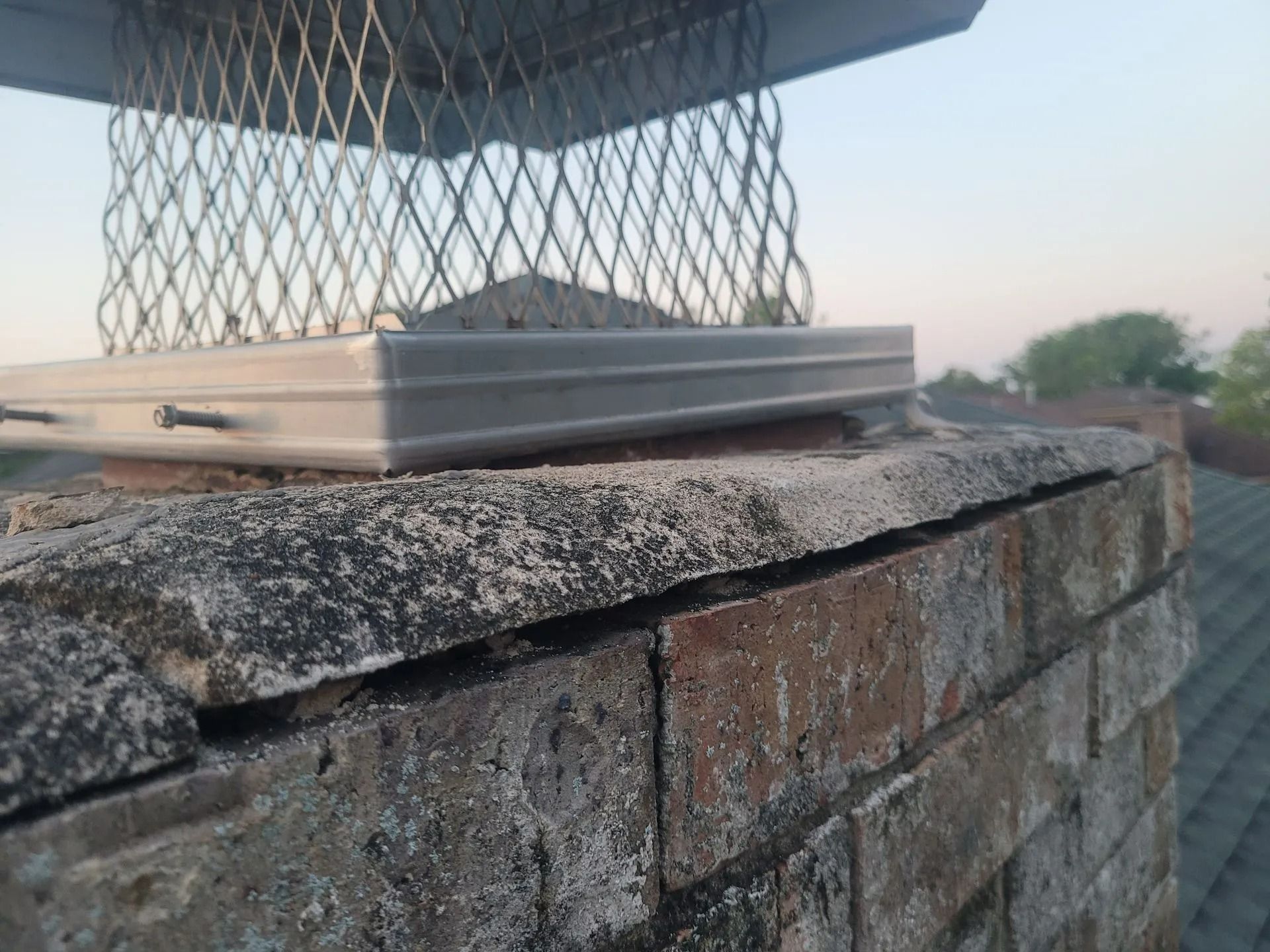 Chimney with a metal cap and mesh screen, showing brick damage and white residue.