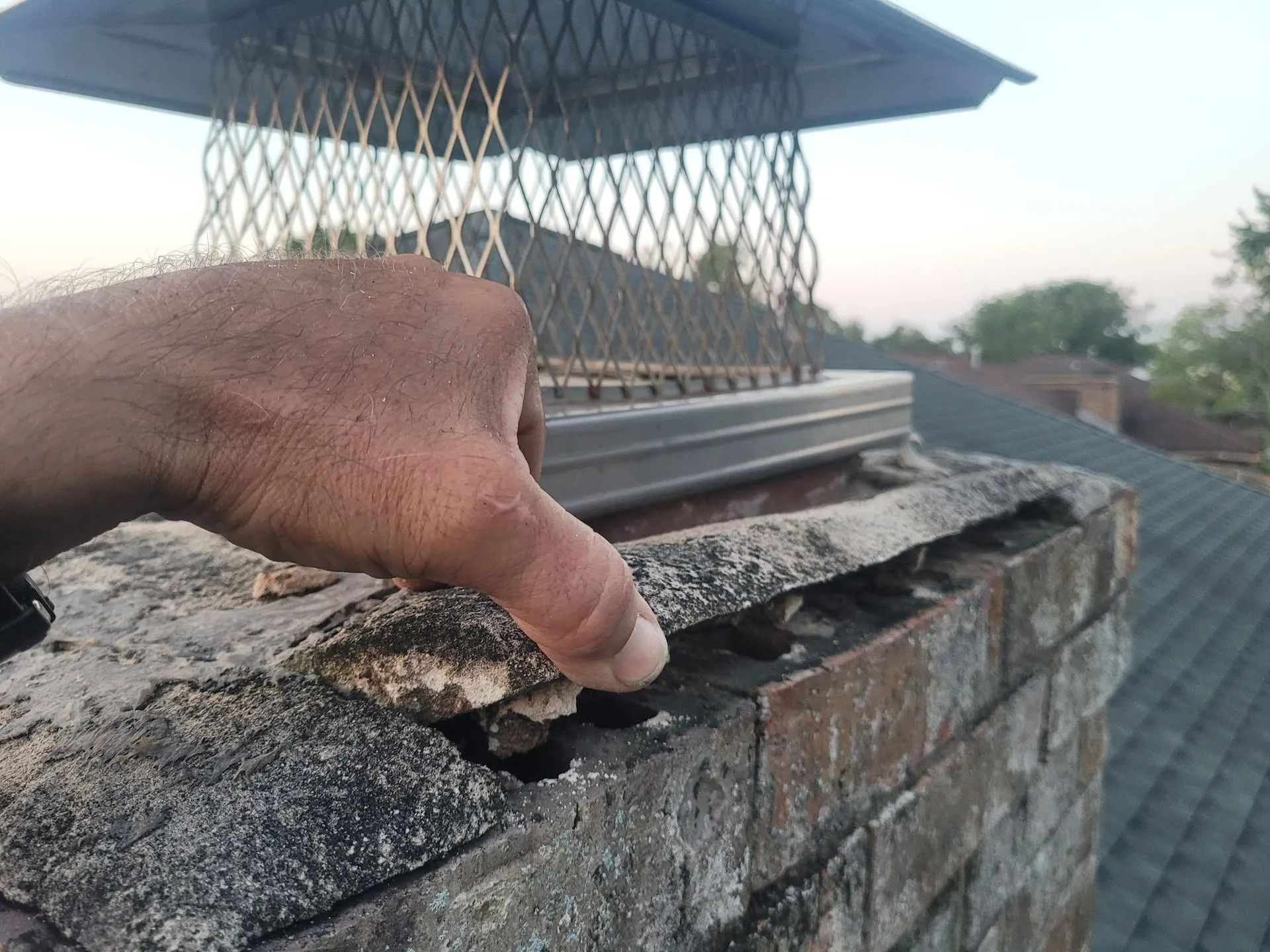 Hand touching a crumbling chimney cap with mesh screen and roof in background.