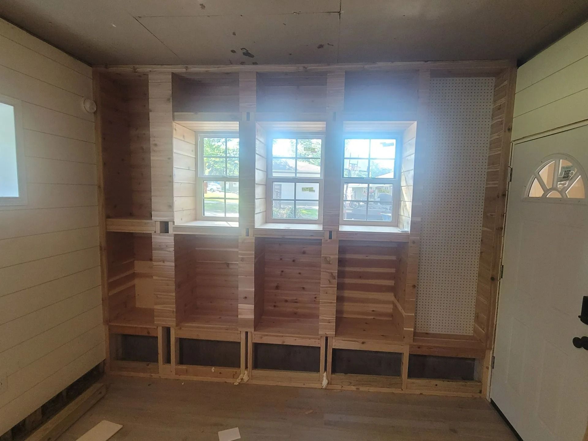 Wooden built-in storage and seating unit under three windows in a room with a door and unfinished walls.
