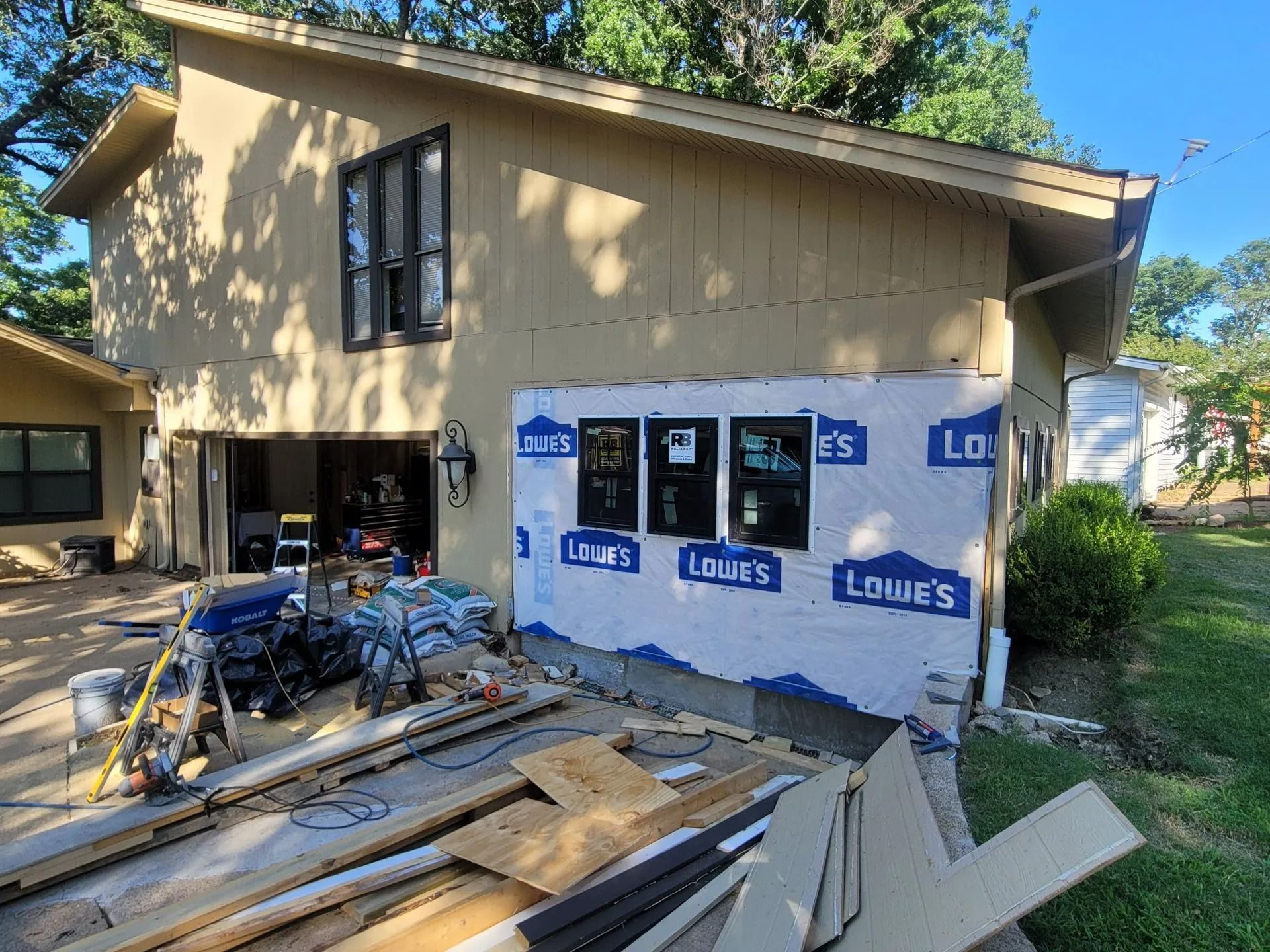 House exterior under construction, with new windows installed and covered in blue Lowe's wrap.