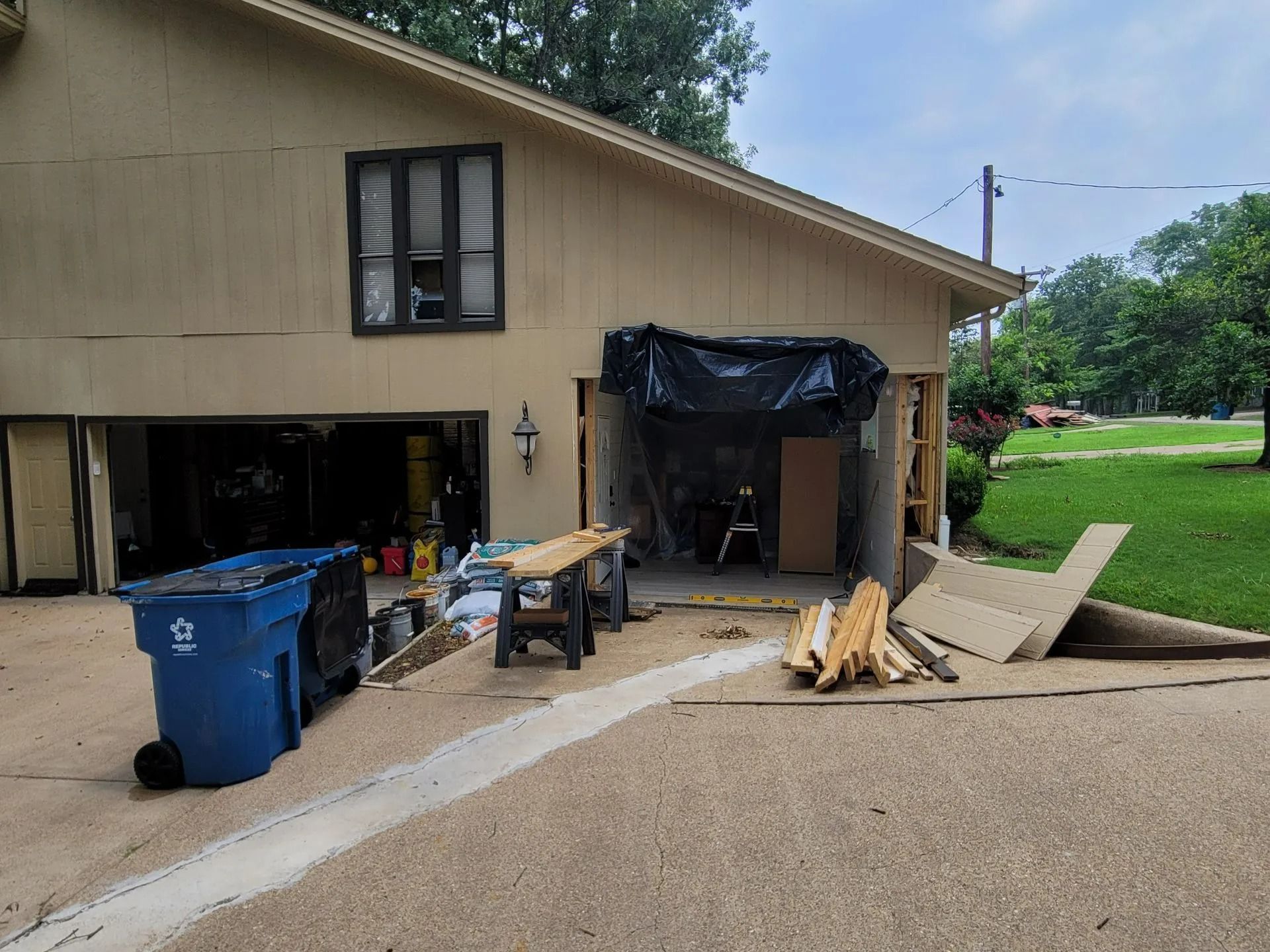 Garage undergoing renovation; one door open, debris and tools visible. Exterior walls are beige, driveway and lawn are visible.
