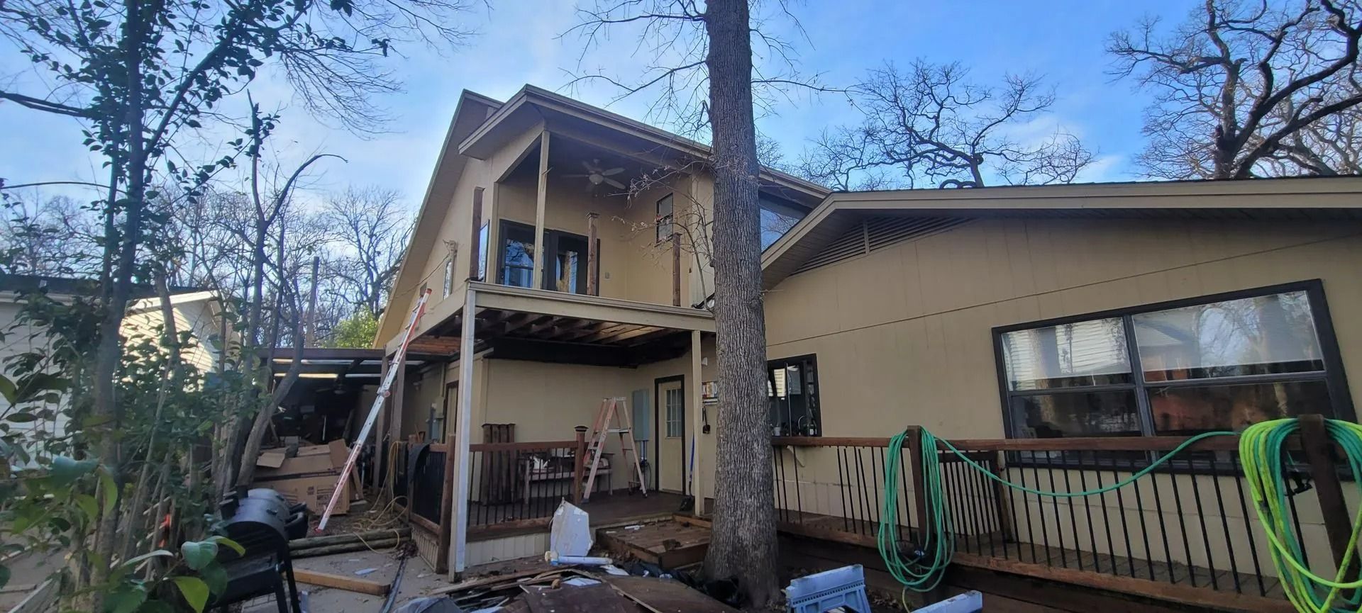 Two-story house with decks, tan siding, and a tree growing beside it. Cloudy sky.