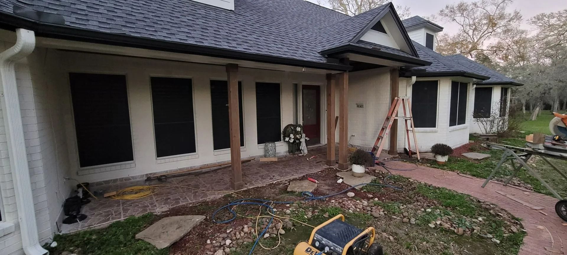 Exterior of a house with black shutters, a red brick walkway, and construction materials.