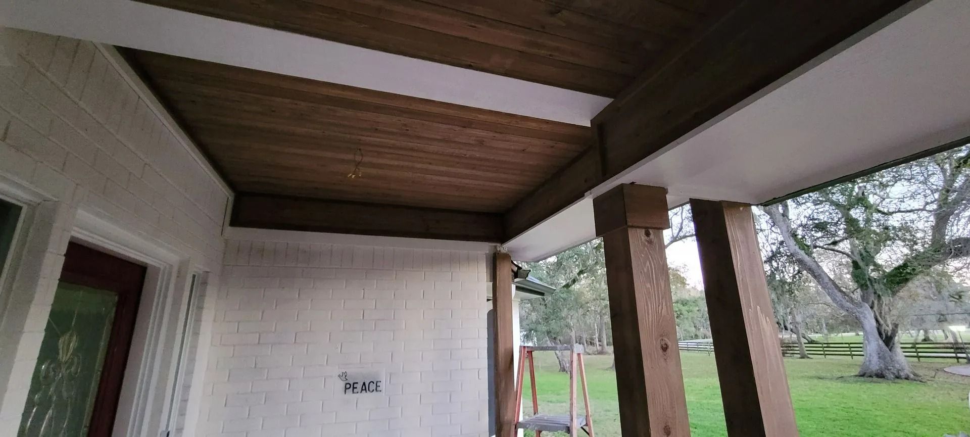 Porch ceiling with white trim and stained wood. Brick wall and wooden pillars, grass and trees in background.