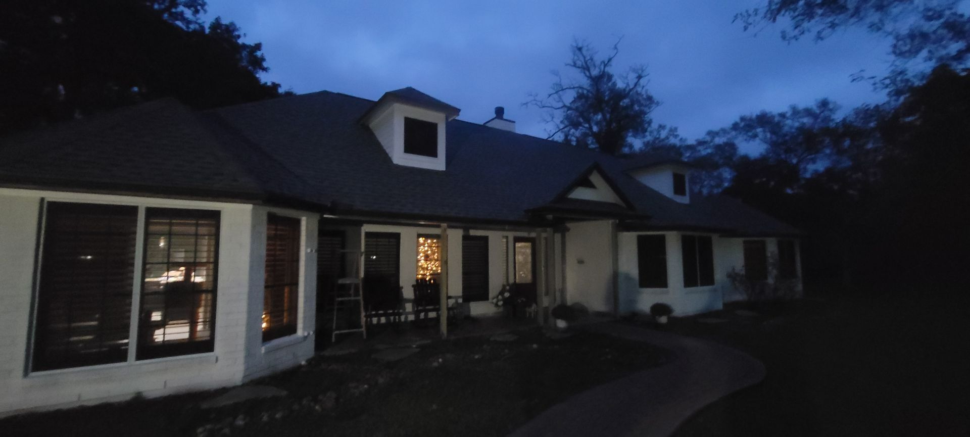 A white house at dusk with lit windows, a path, and trees.