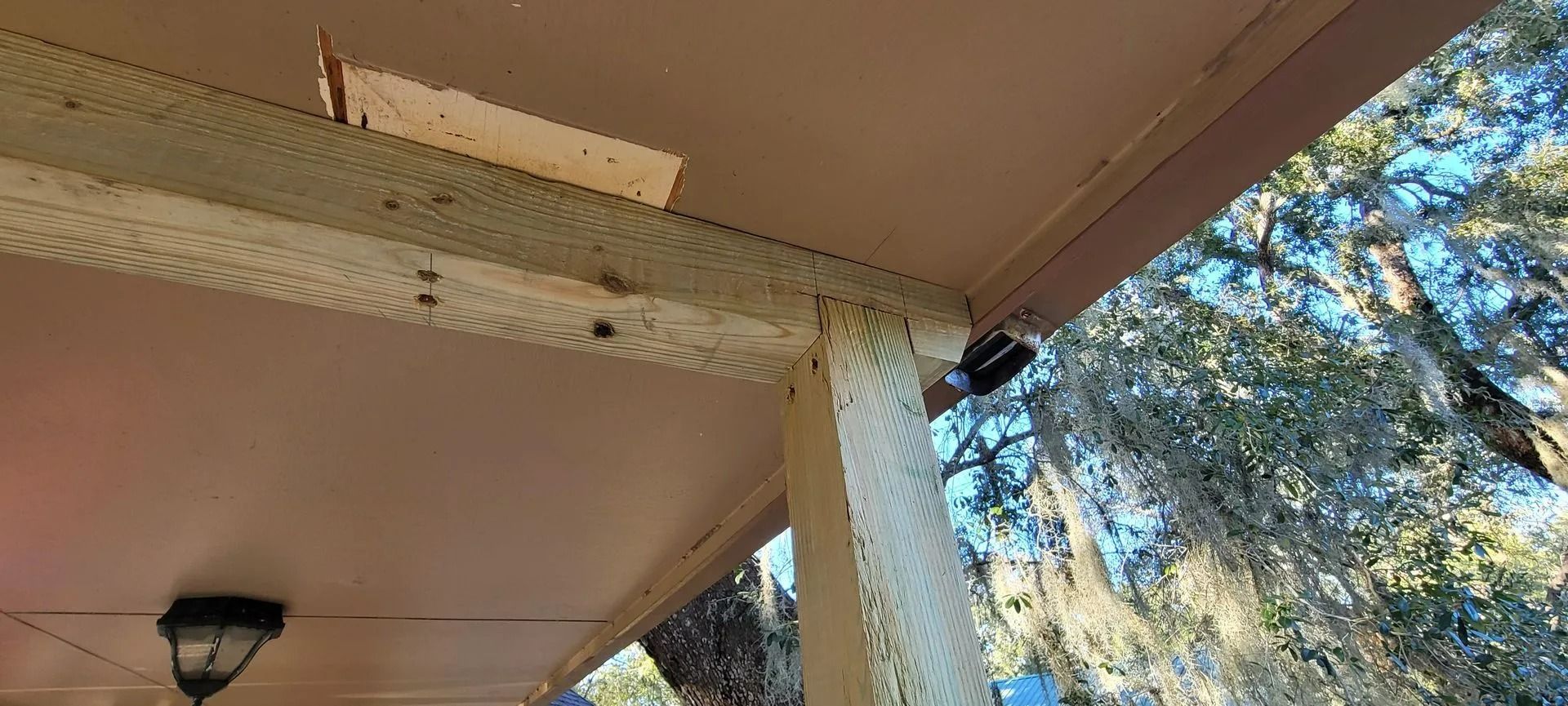 View from below of a porch roof, column, and ceiling fan. The porch overlooks trees and sky.