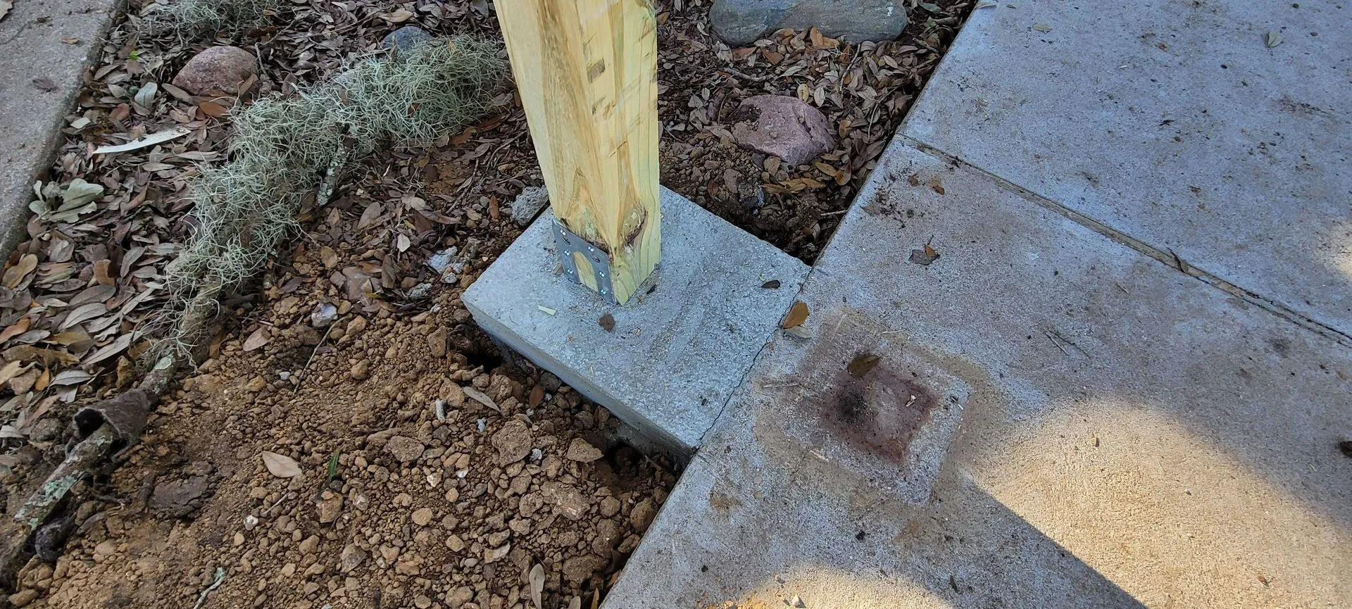 Wooden post attached to a concrete base near a concrete walkway and dirt.