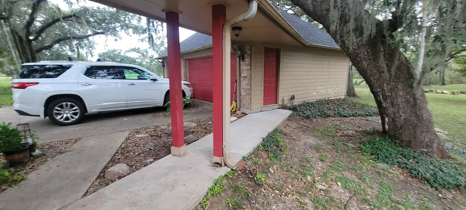 White SUV parked under a carport. Red supports and door frame on a beige building with a tree.