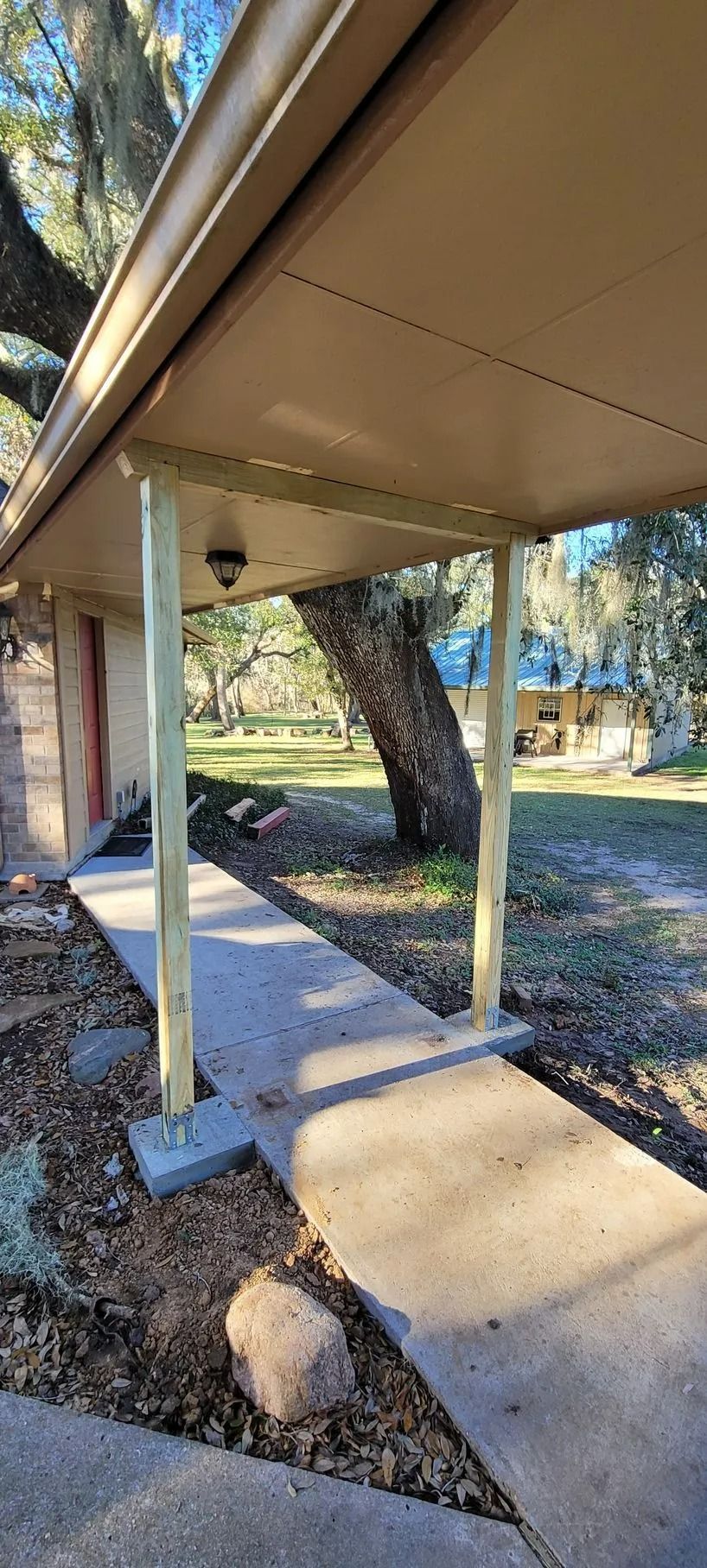 Concrete walkway with a covered porch supported by wooden posts. A large tree stands in the background.
