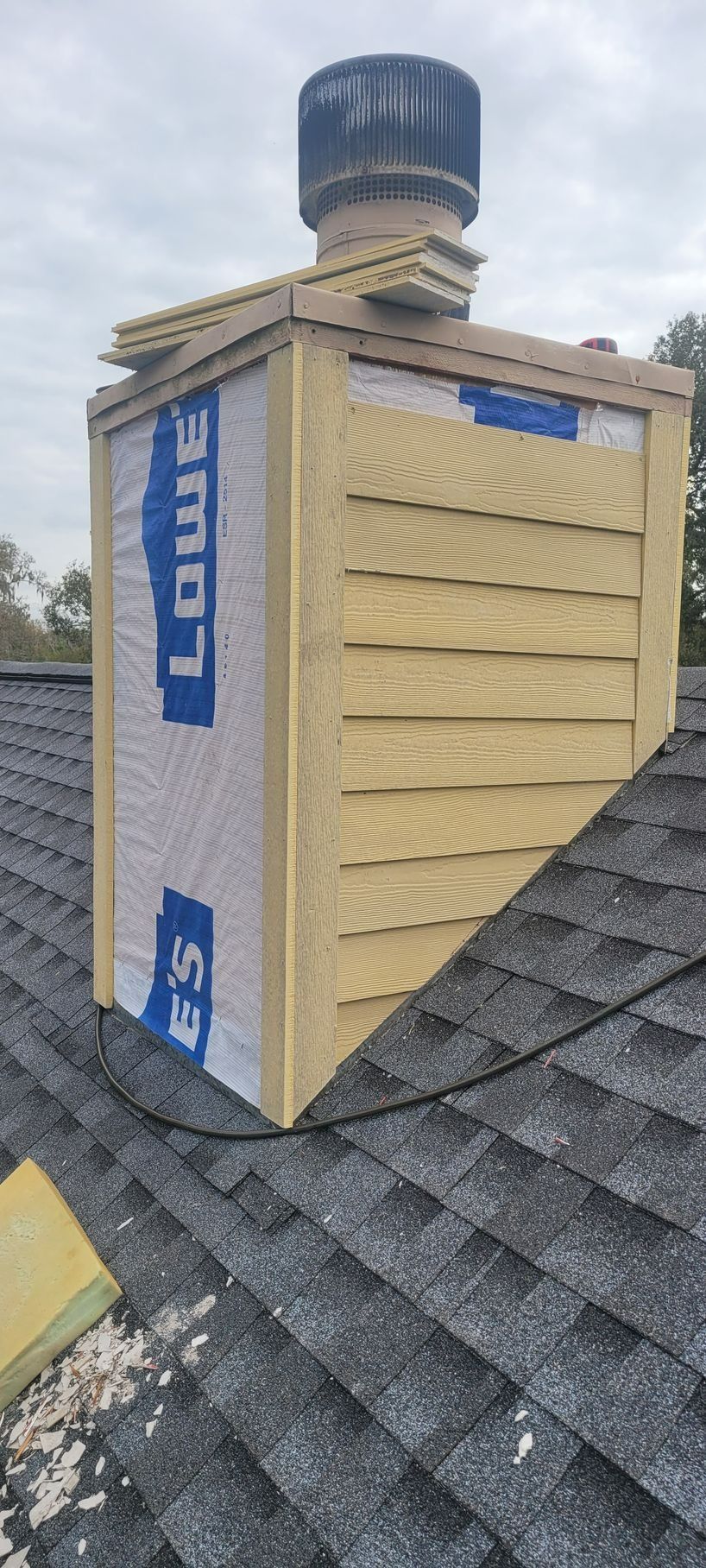 Chimney on a roof. Wooden frame with a Lowe's insulation wrap. Top has a metal cap, shingles around base.