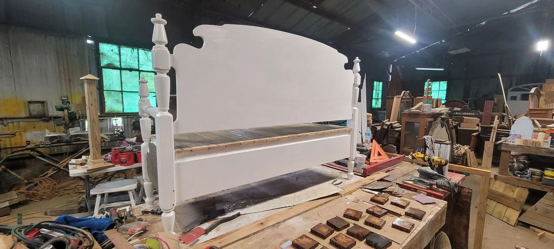 A white bed frame under construction in a workshop. Tools and materials surround the frame, window in background.