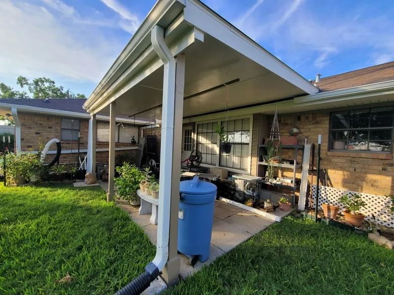 Backyard with patio cover, brick house, green grass, blue sky, and a blue trash can.