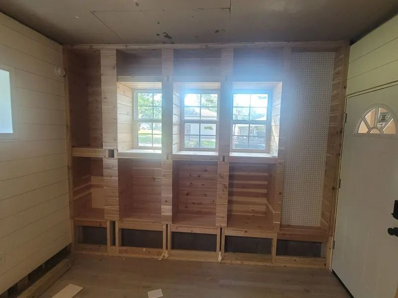 Wooden built-in storage unit with cubbies, benches, and windows under construction in a room.