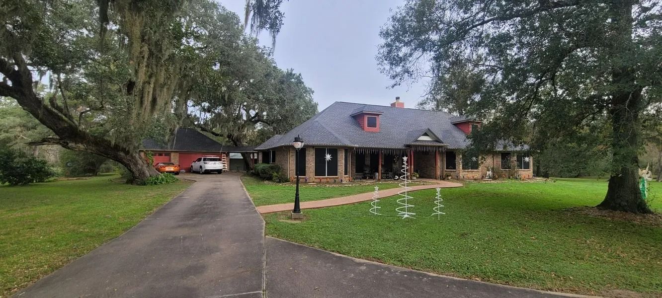 A house with a long driveway. Trees frame the brick exterior and green lawn.