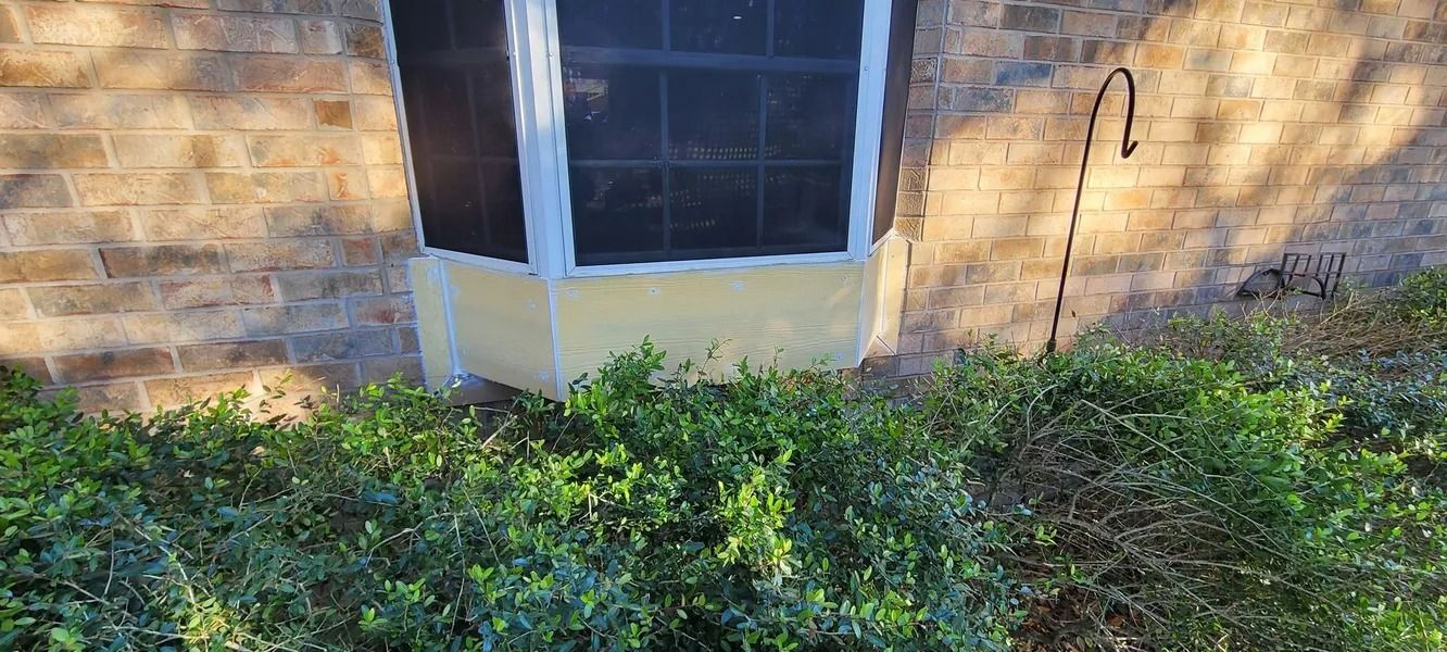 A brick building with a window, yellow trim, and a curved metal hook next to a bush.