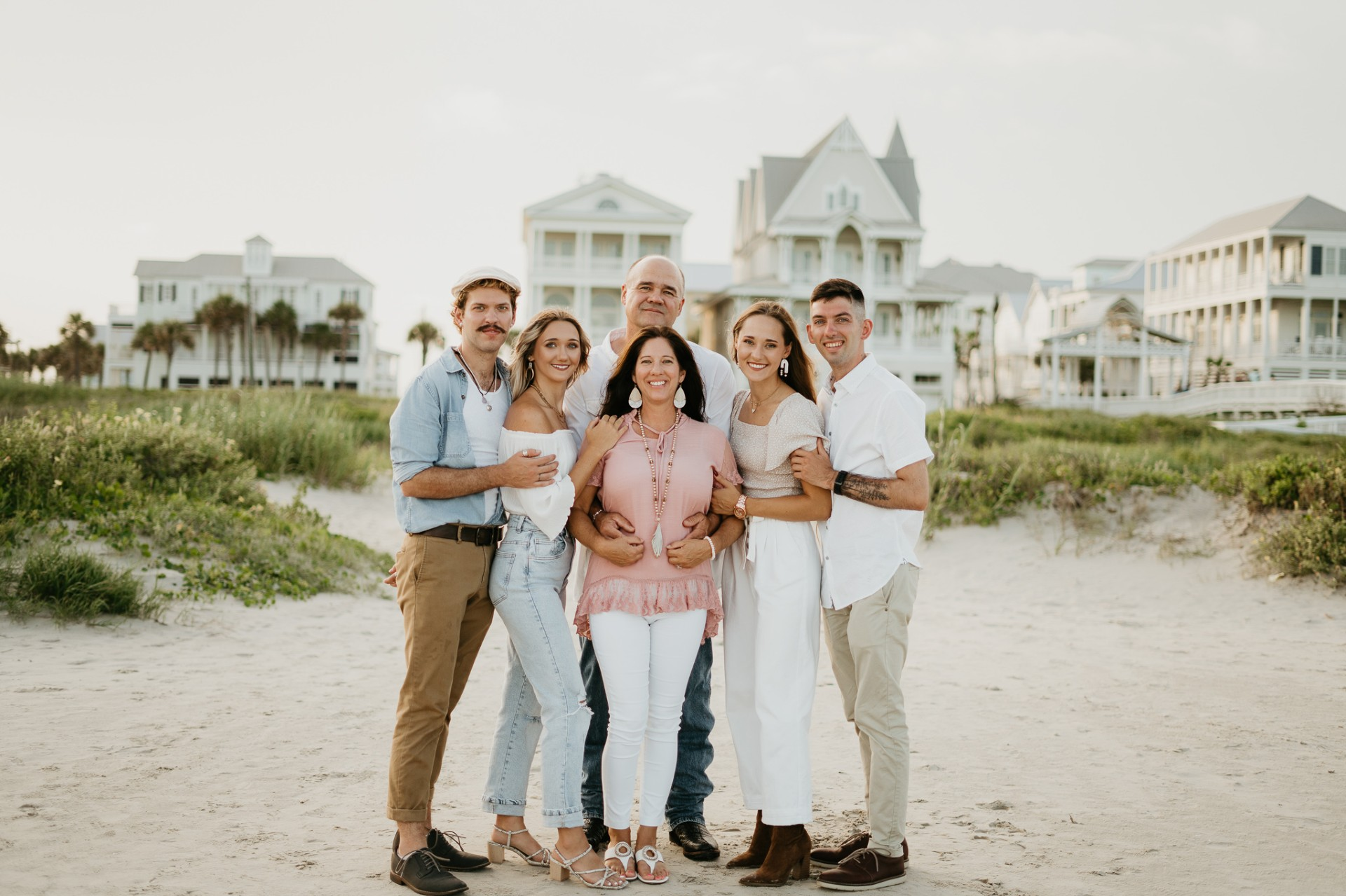 Family on a beach, smiling and posing in front of white buildings; sand and greenery in foreground.
