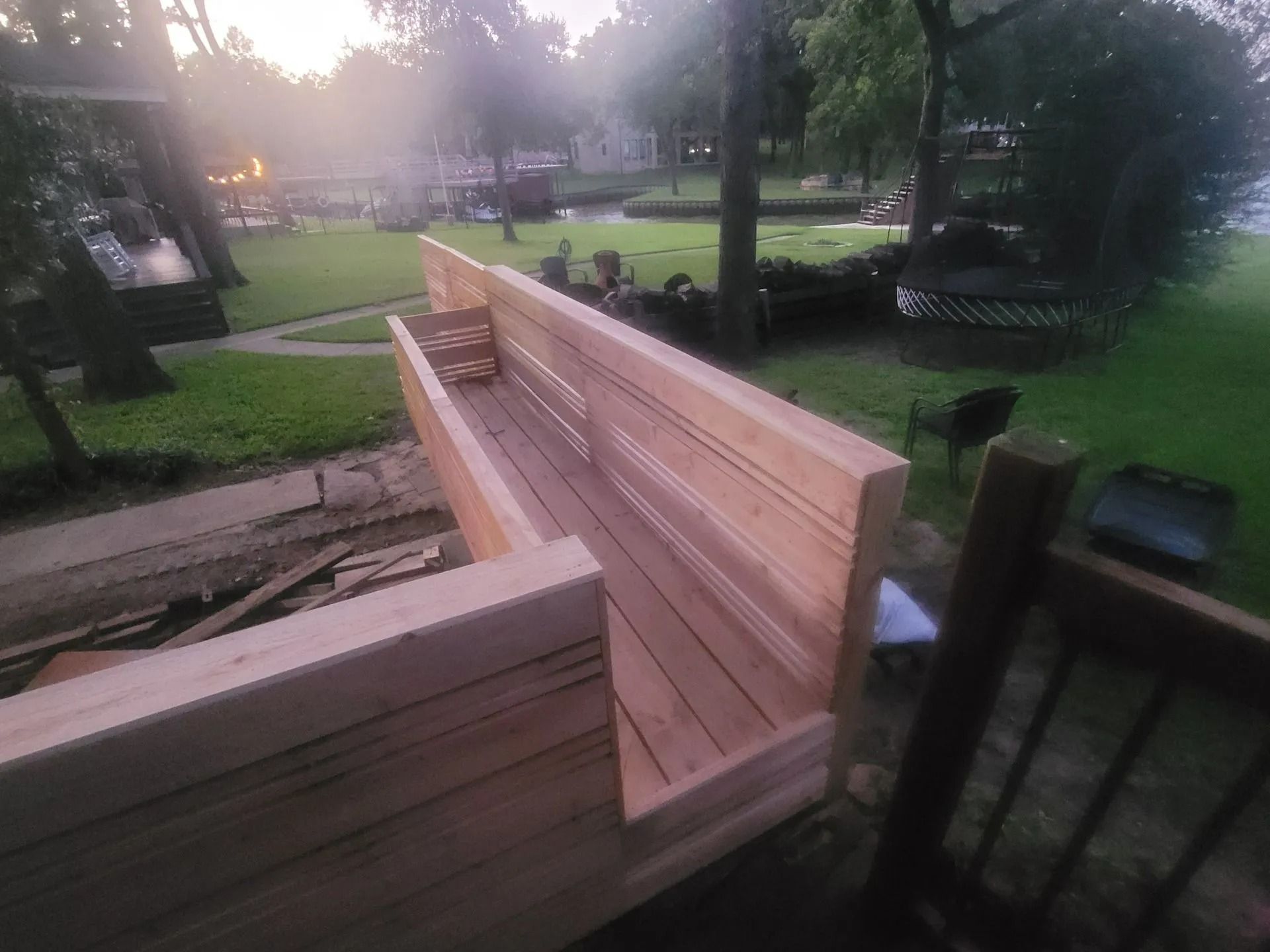Wooden deck under construction, overlooking a grassy area, trees, and water. Cloudy sky.