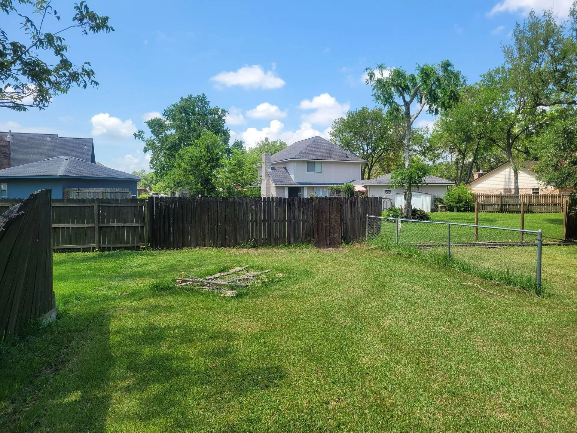 Green backyard with wooden fence, trees, and houses under a partly cloudy sky.