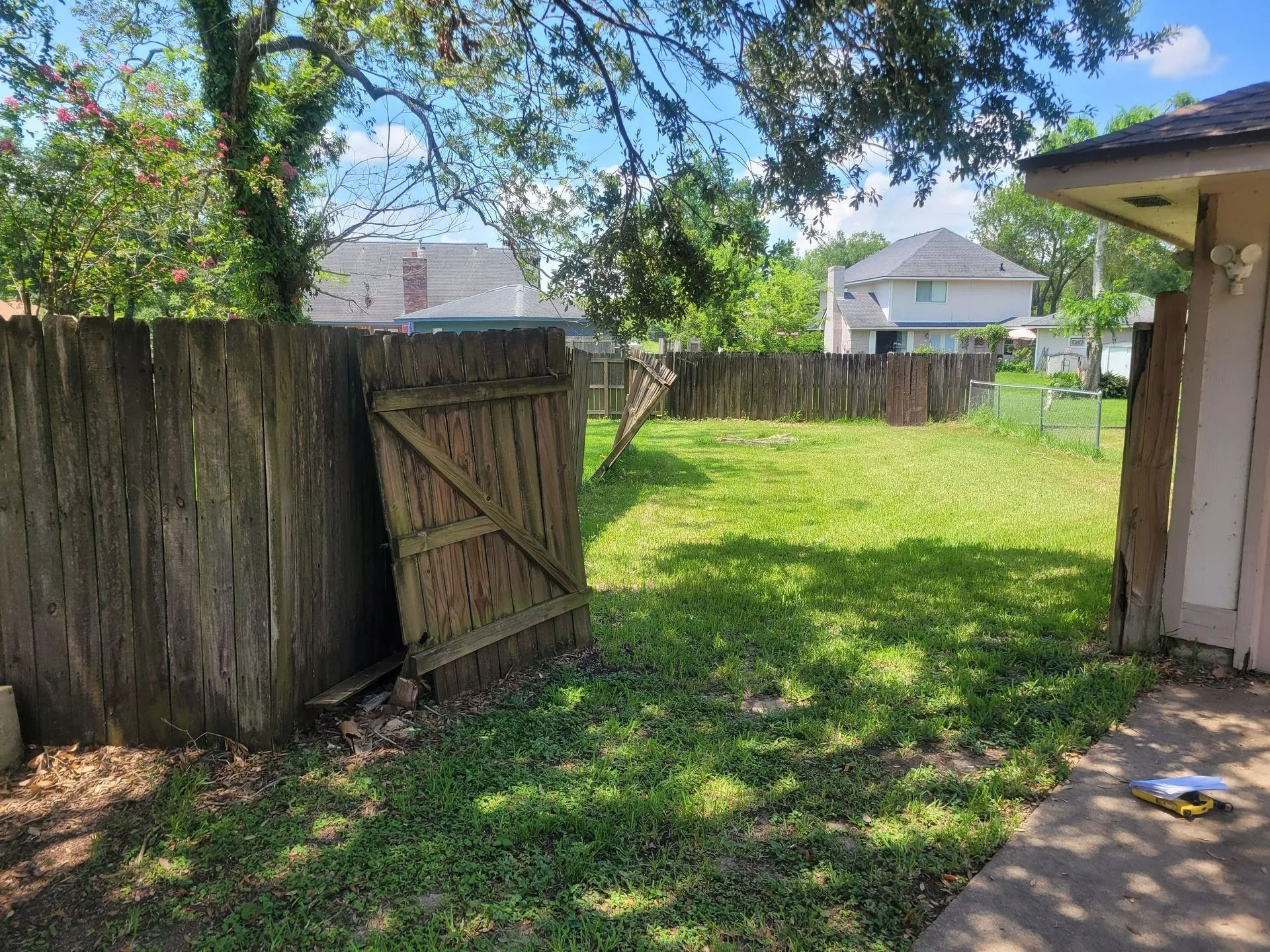 Backyard with a wooden fence and gate, green grass, and houses in the distance.