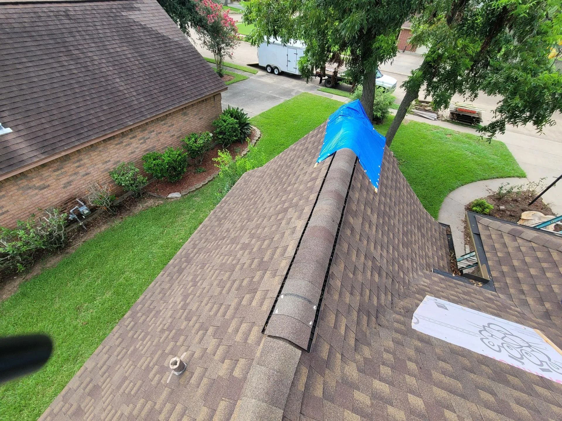 Overhead view of a house roof with a blue tarp covering a section, brown shingles, and green grass.