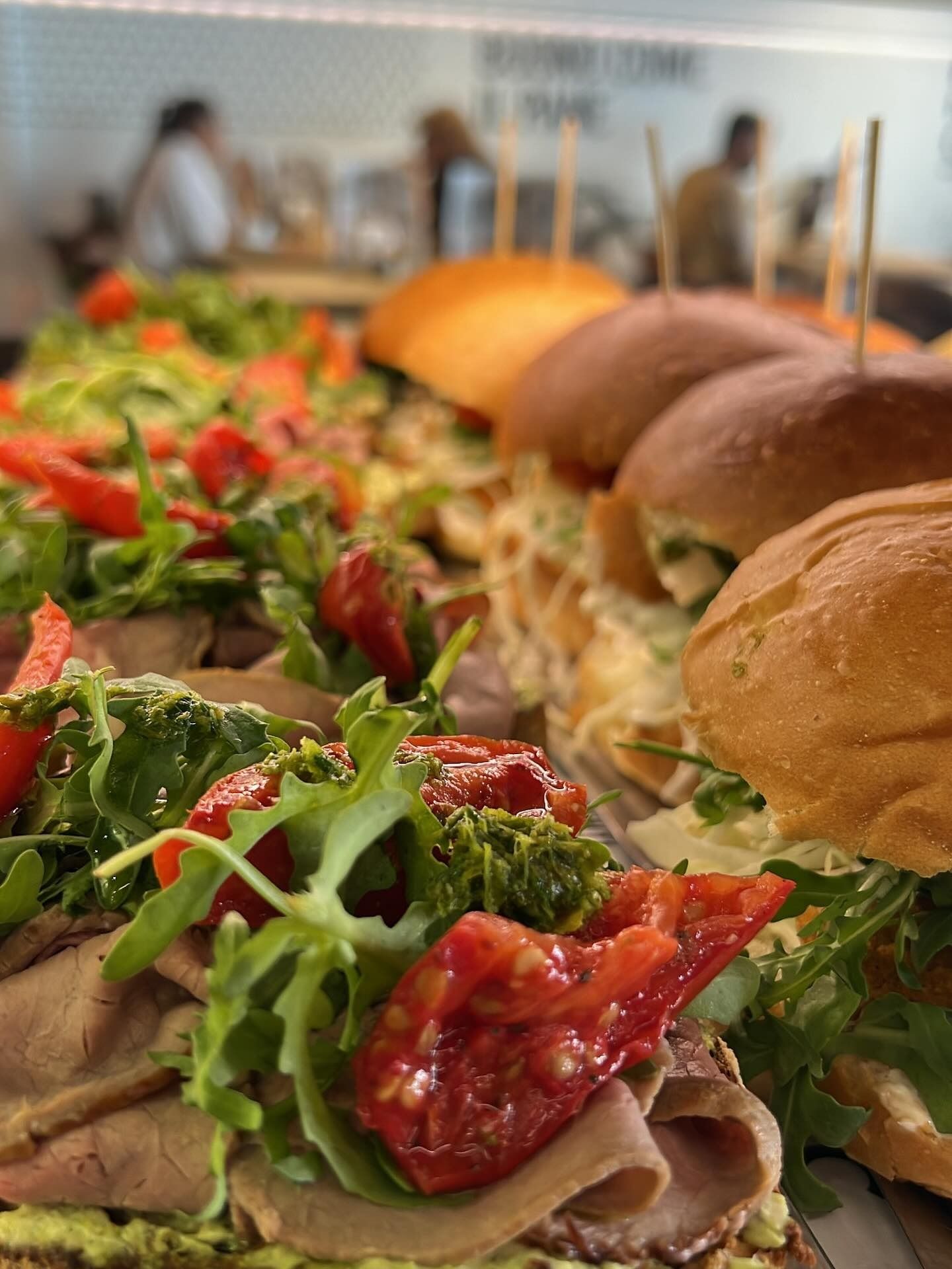 Primo piano di panini esposti su un espositore. Condimenti a base di rucola, pomodori e pesto. Le persone sono sfocate sullo sfondo.
