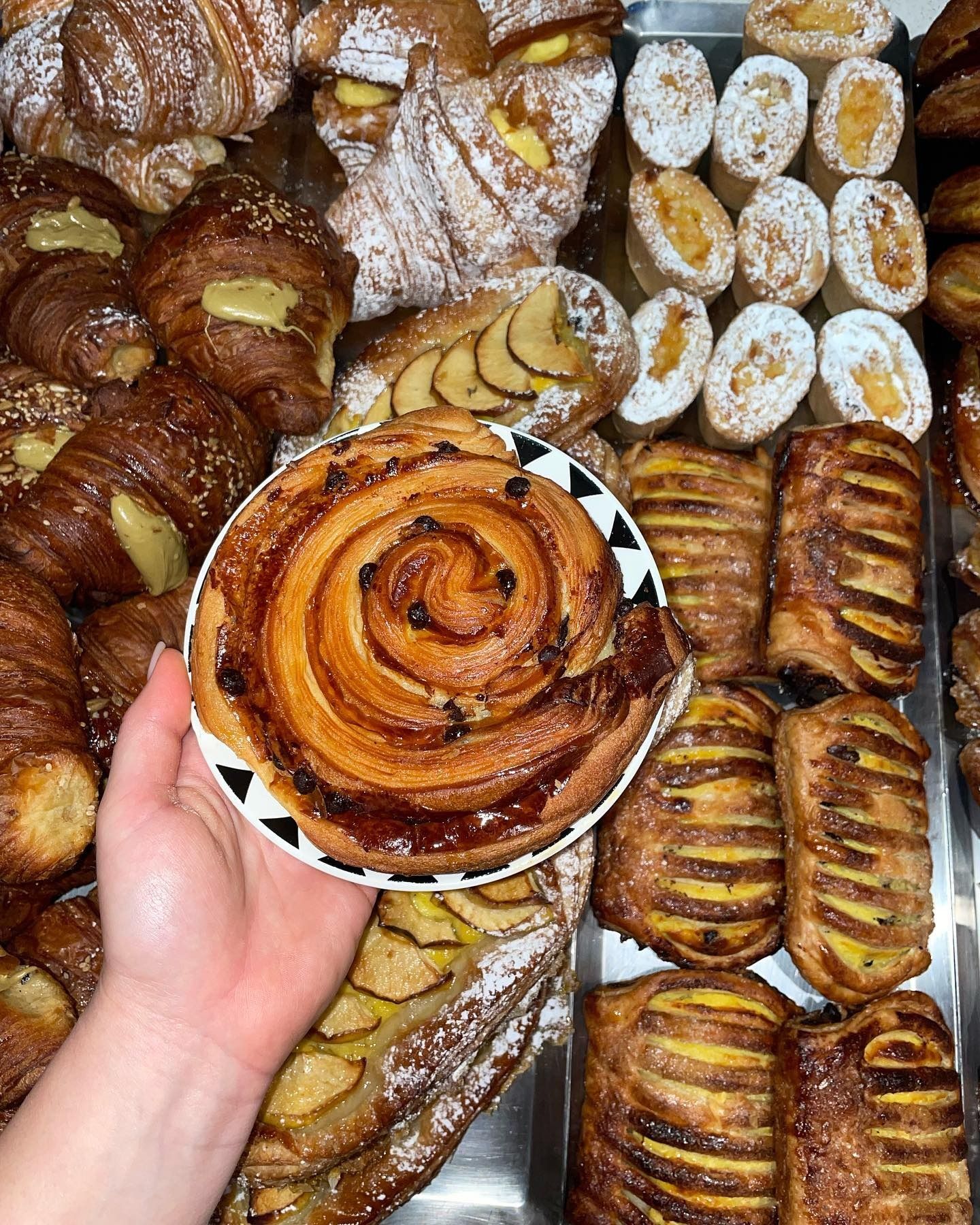 Mano che tiene un dolce rotondo con gocce di cioccolato, circondato da prodotti da forno assortiti, su un vassoio di metallo.