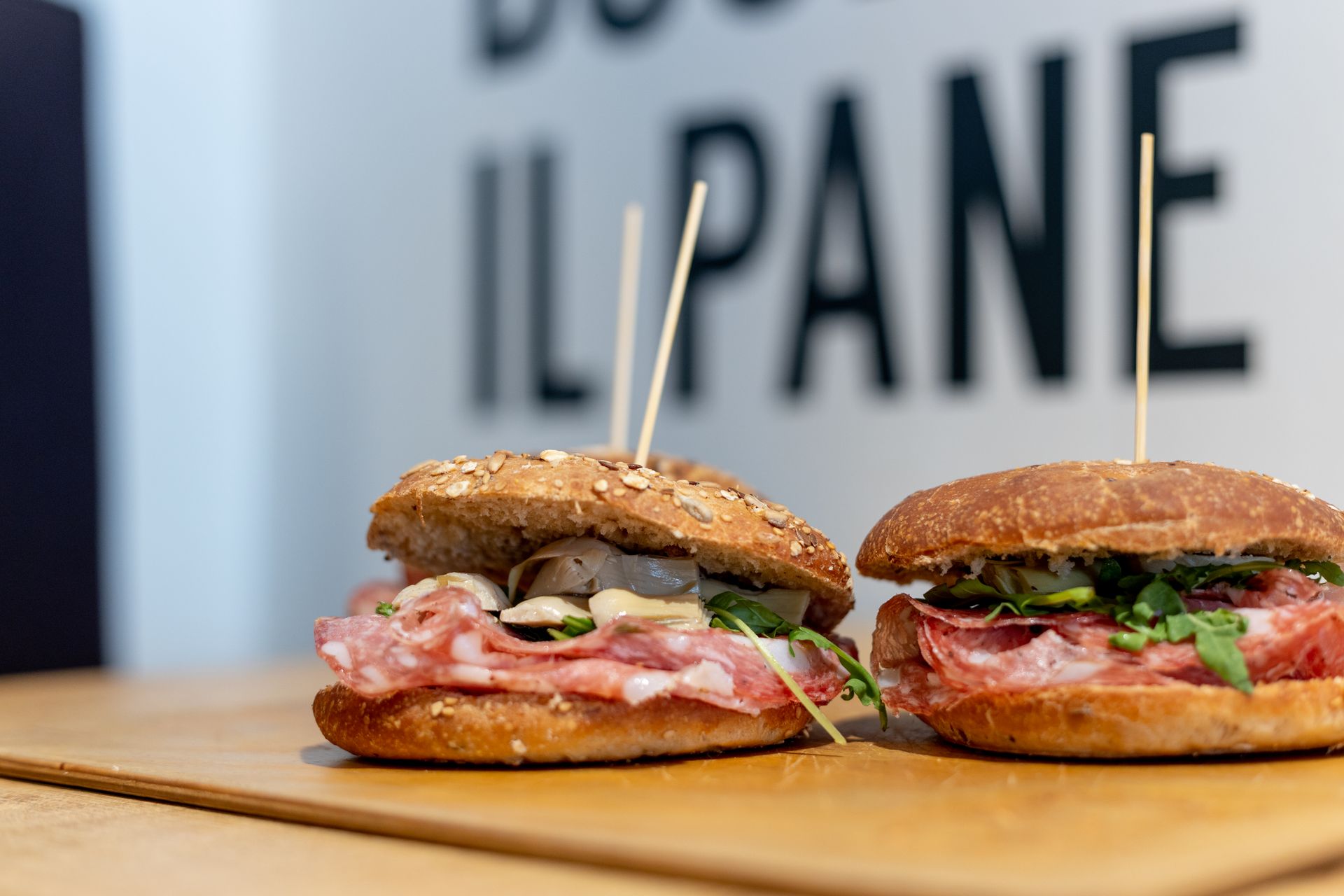 Due panini su un tagliere di legno, con salame, rucola e stuzzicadenti, contro una parete bianca e sfocata.