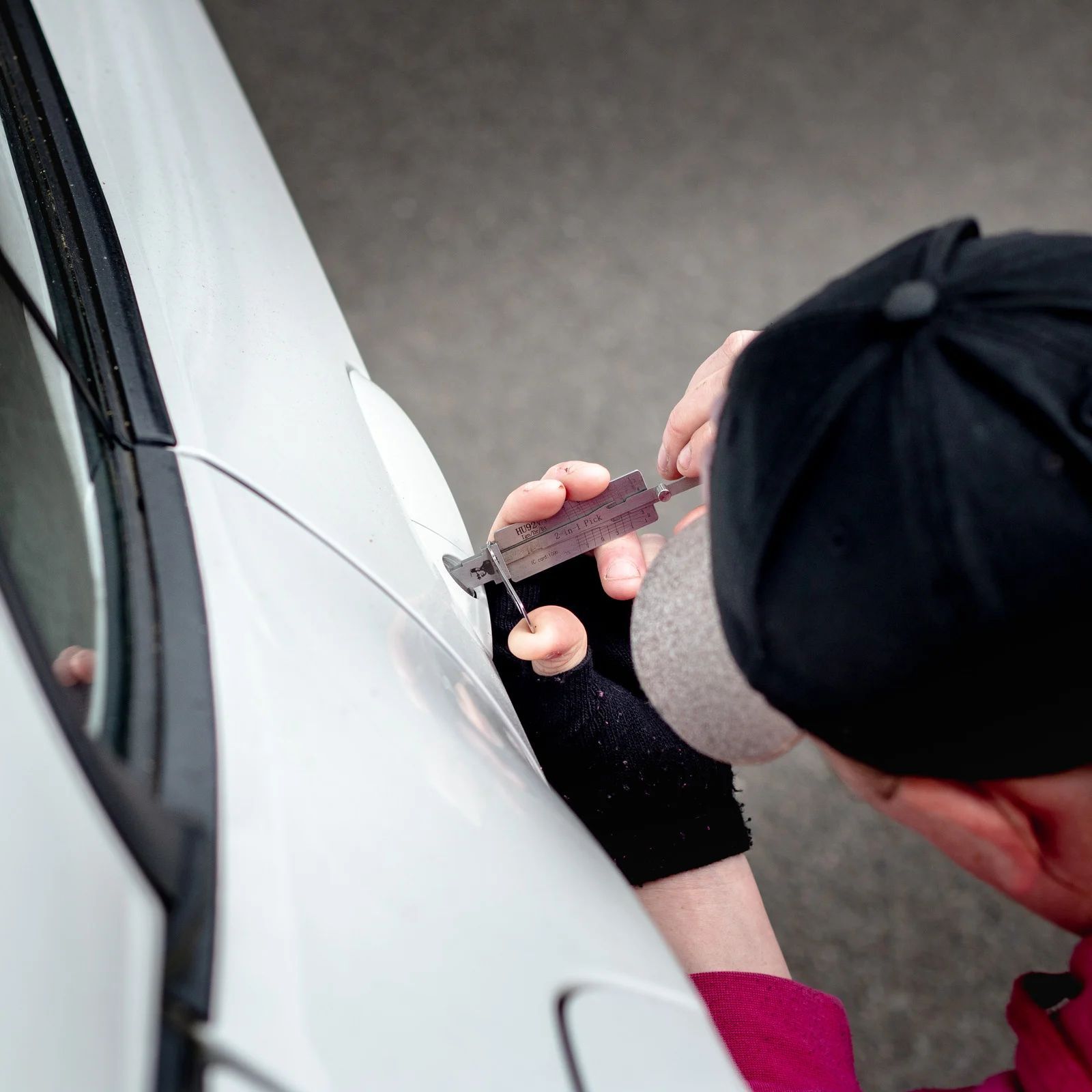Person in a cap uses lock picks on a white car door. They are close up, with tools in hand.