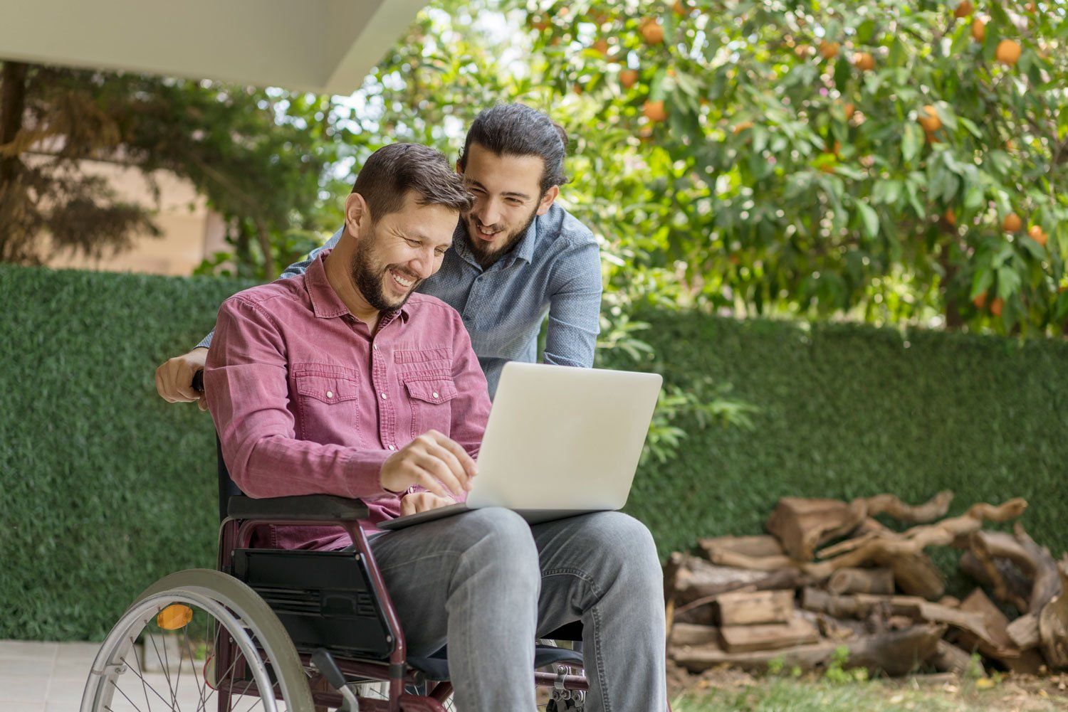 Two young men, one in a wheelchair, the other standing, enjoy a video call on a laptop in the backyard.