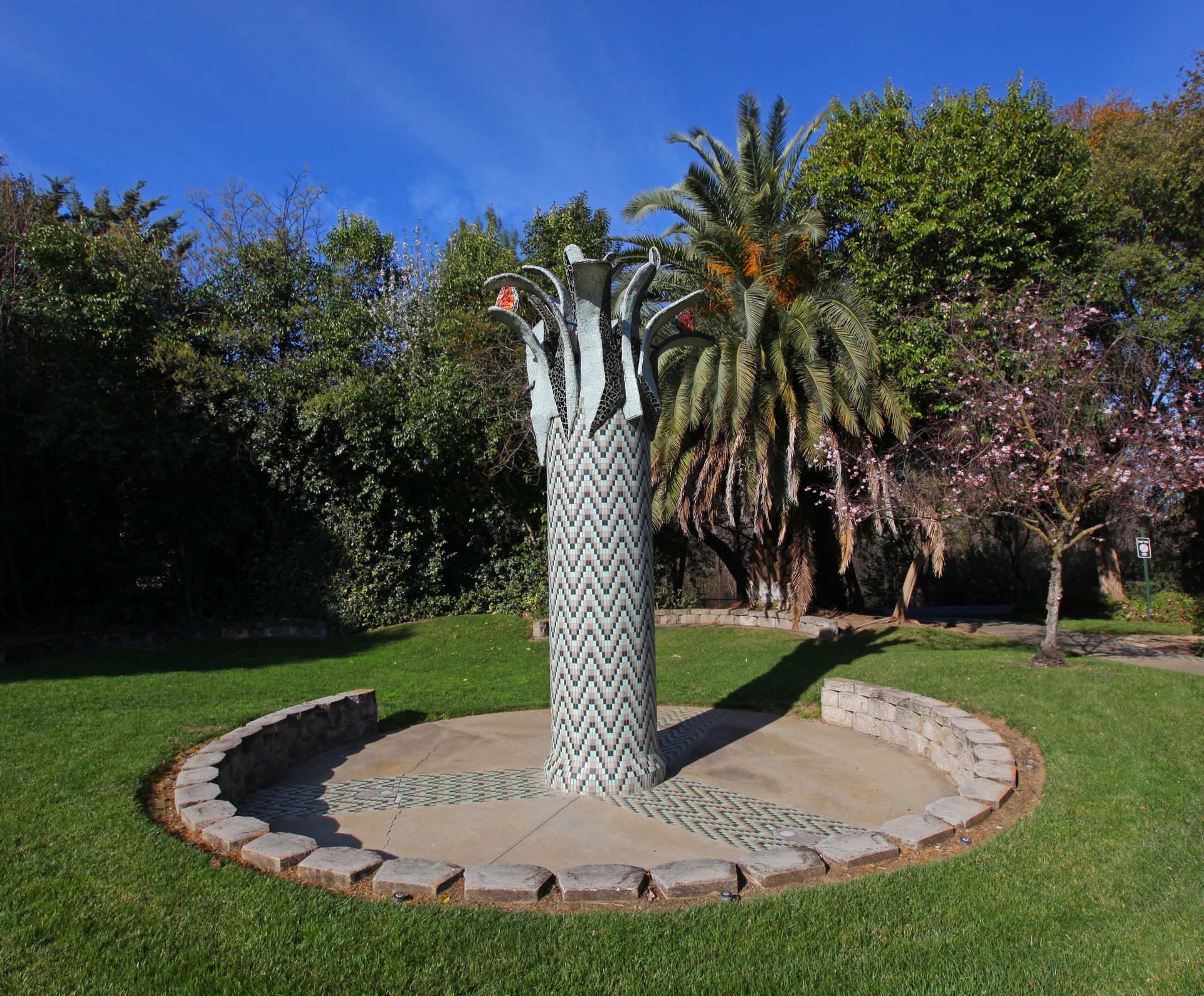 Sculpture with a textured column and leafy top in a park, surrounded by greenery and a circular stone border.