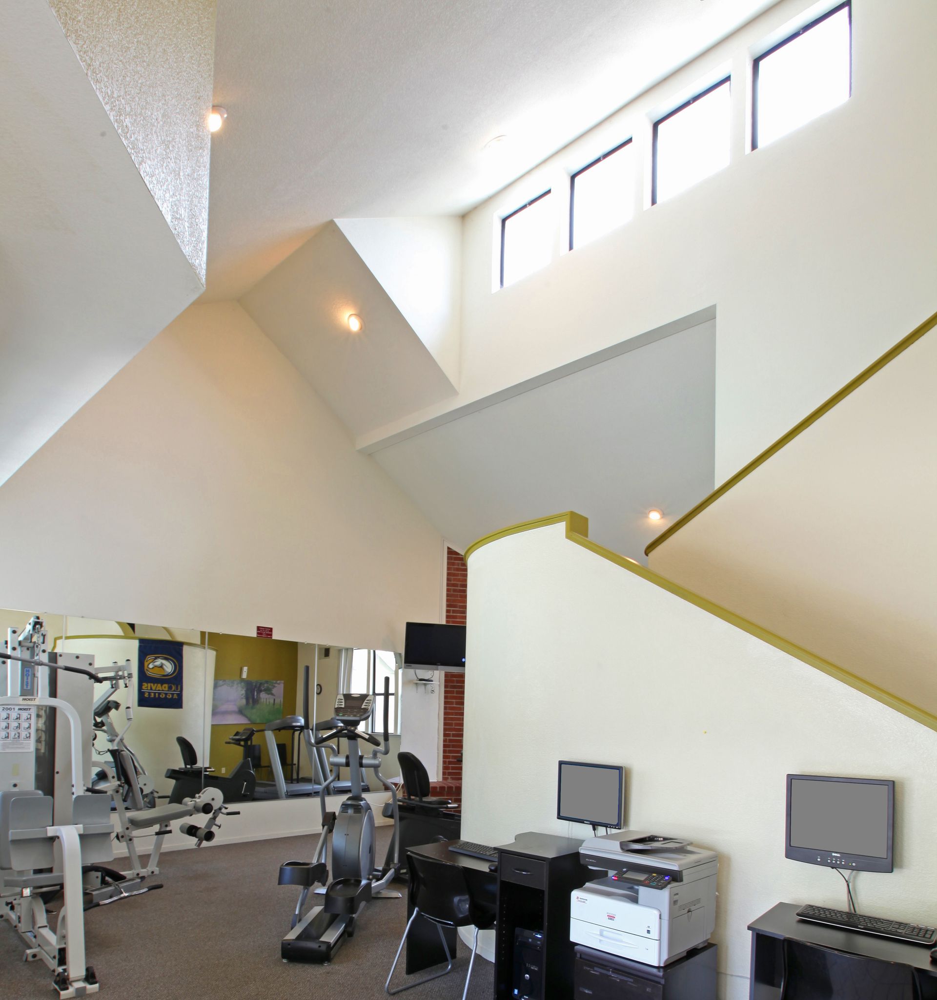 Gym with exercise machines, computer desk, high ceilings, and windows.