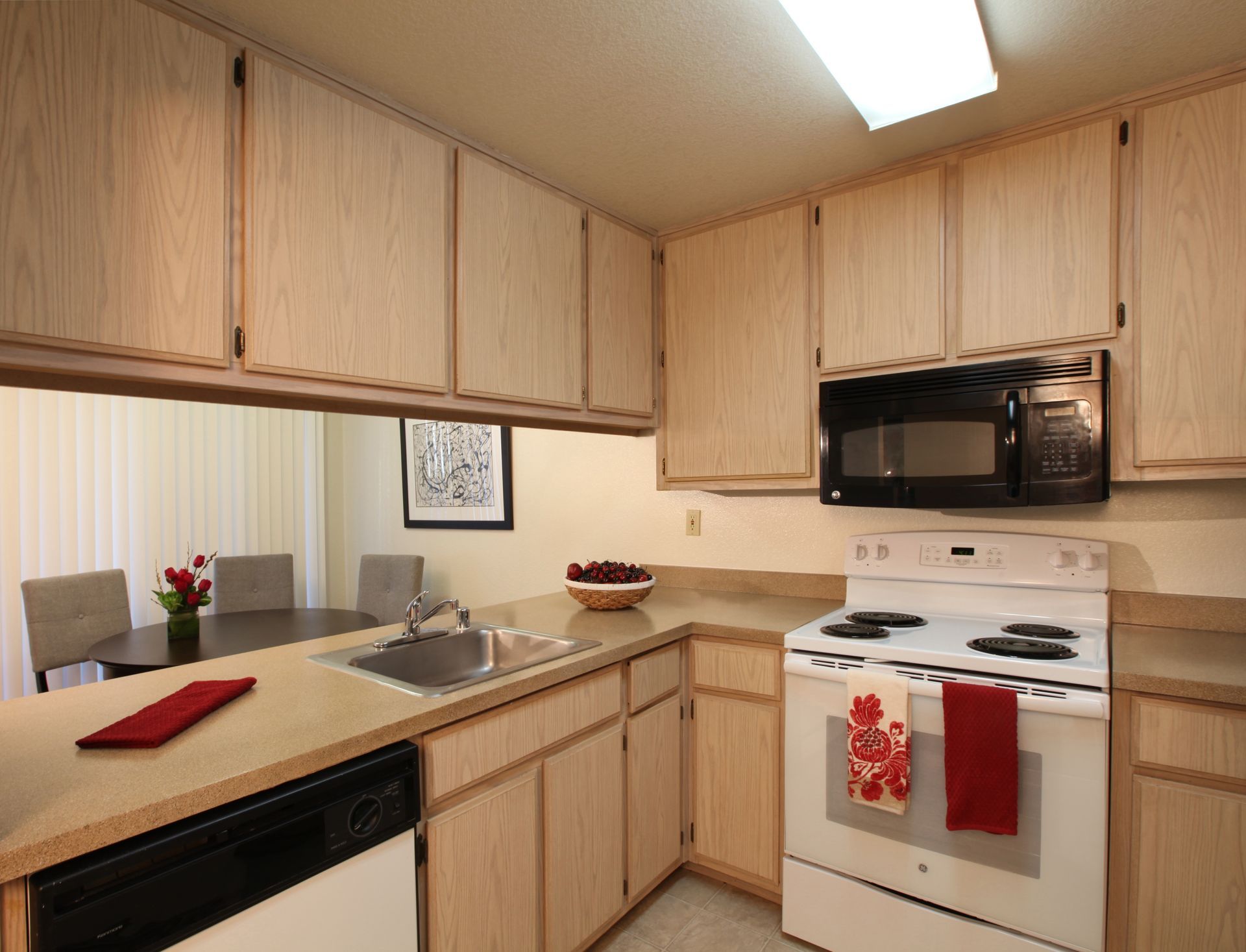 Kitchen with light wood cabinets, appliances, and a view of a dining area.