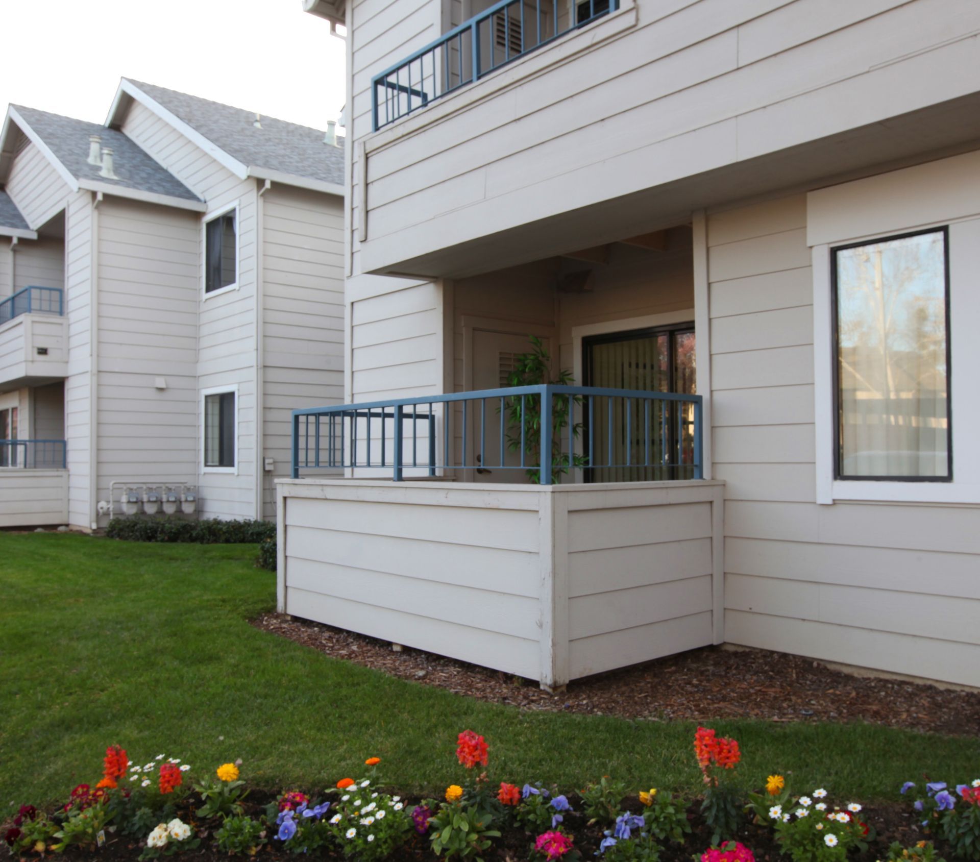 Apartment building with beige siding, blue balcony railings, and a flower bed in front of the green lawn.