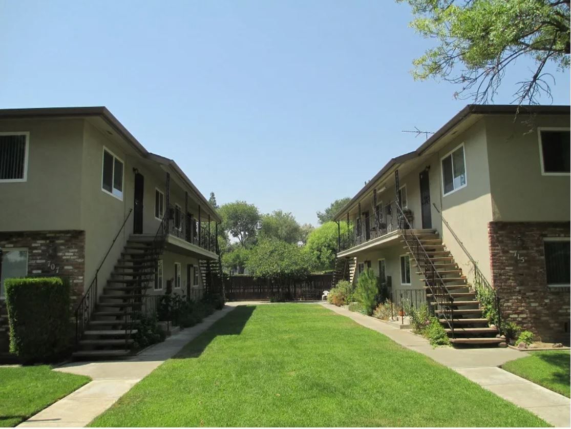 Two-story apartment buildings face each other, with staircases and a grassy courtyard. Beige buildings, blue sky.