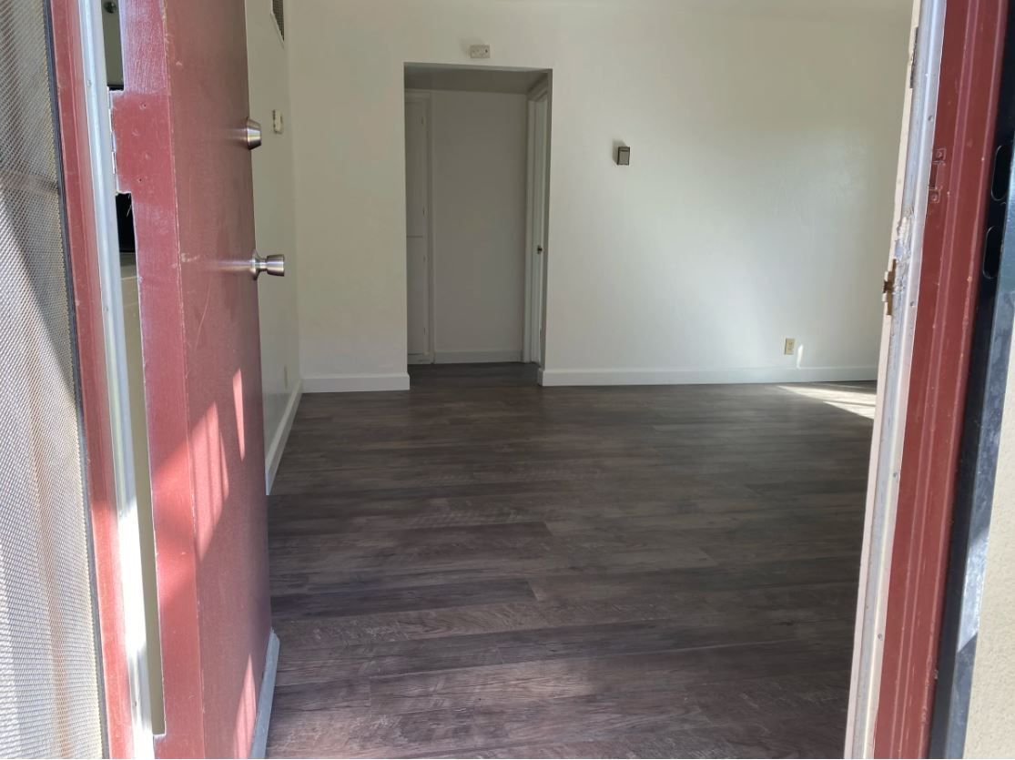 Open doorway into a room with wood-look floors, white walls, and a closed door at the end of a hallway.