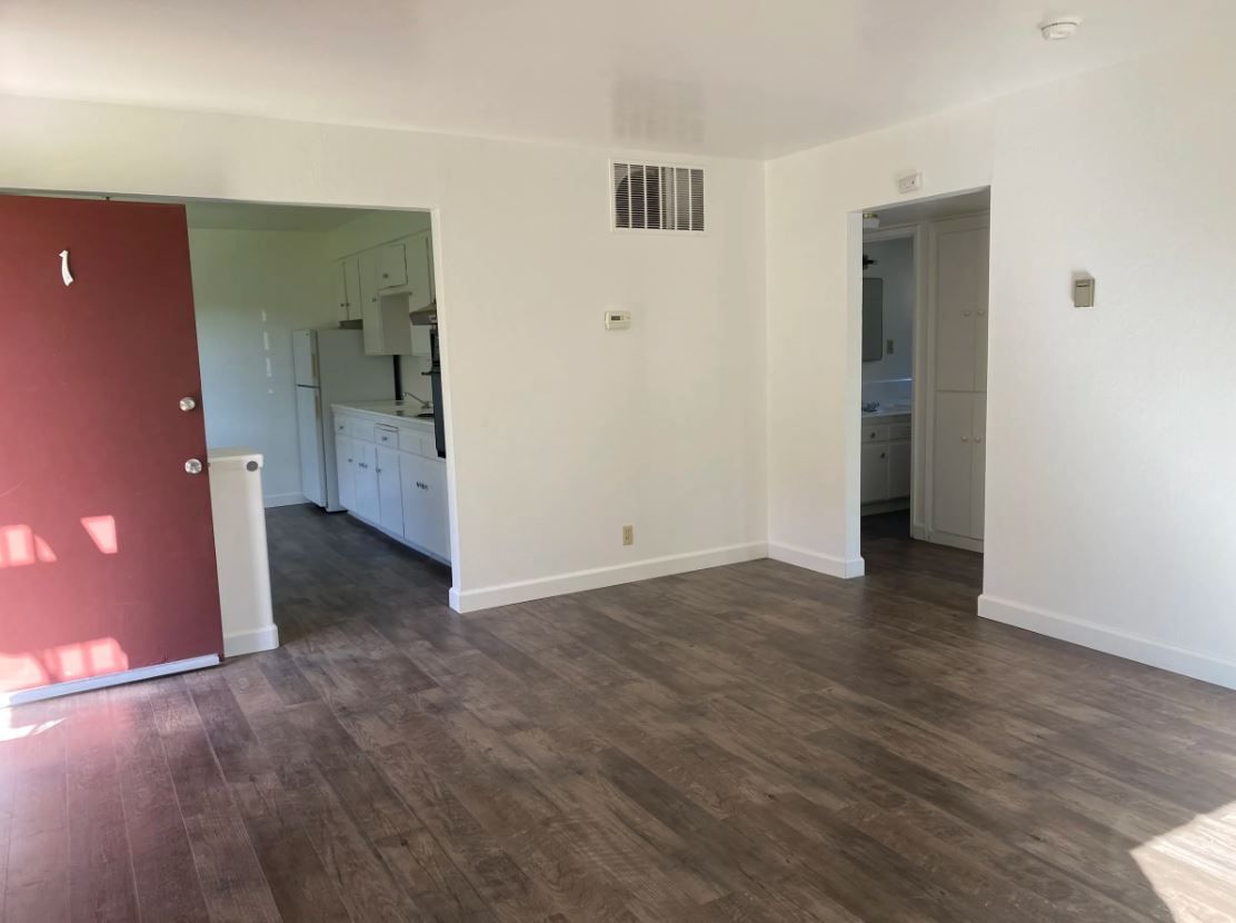 Empty room with wood-look flooring, white walls, and doorways leading to kitchen and bathroom, red door.