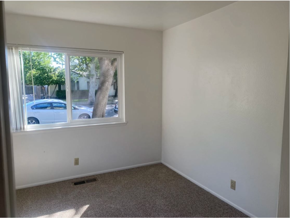 Empty room with a window looking out to a street, light-colored walls, and a patterned carpet.