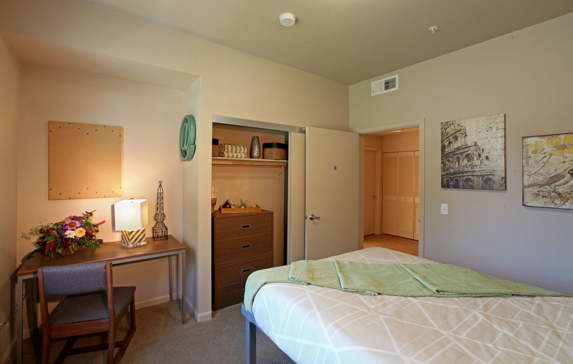 Bedroom with bed, desk, closet, and art. Light-colored walls and a bedspread with a green blanket.