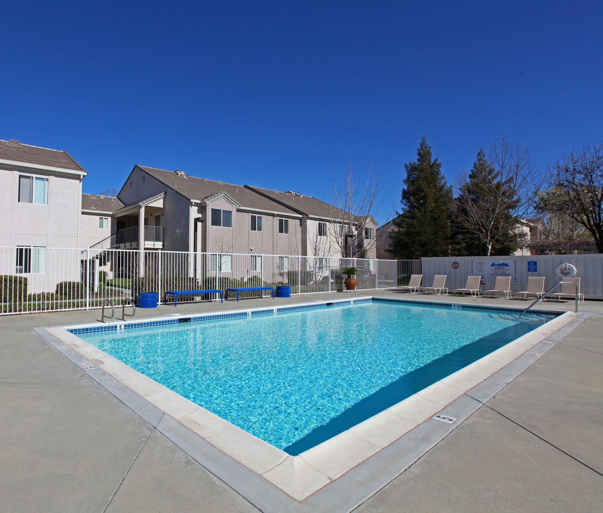 Swimming pool outside apartment buildings on a clear, sunny day. Lounge chairs line the pool deck.