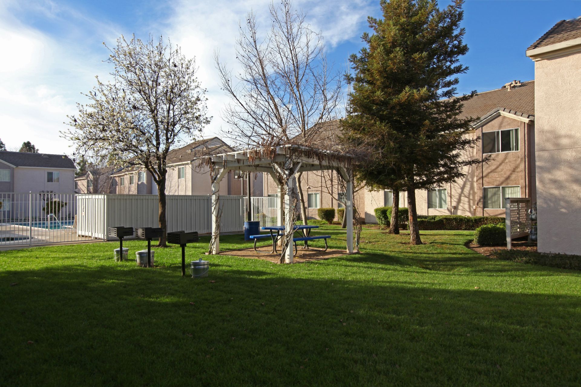 Grassy courtyard with picnic tables, BBQ grills, and a pool, in front of two-story apartment buildings.