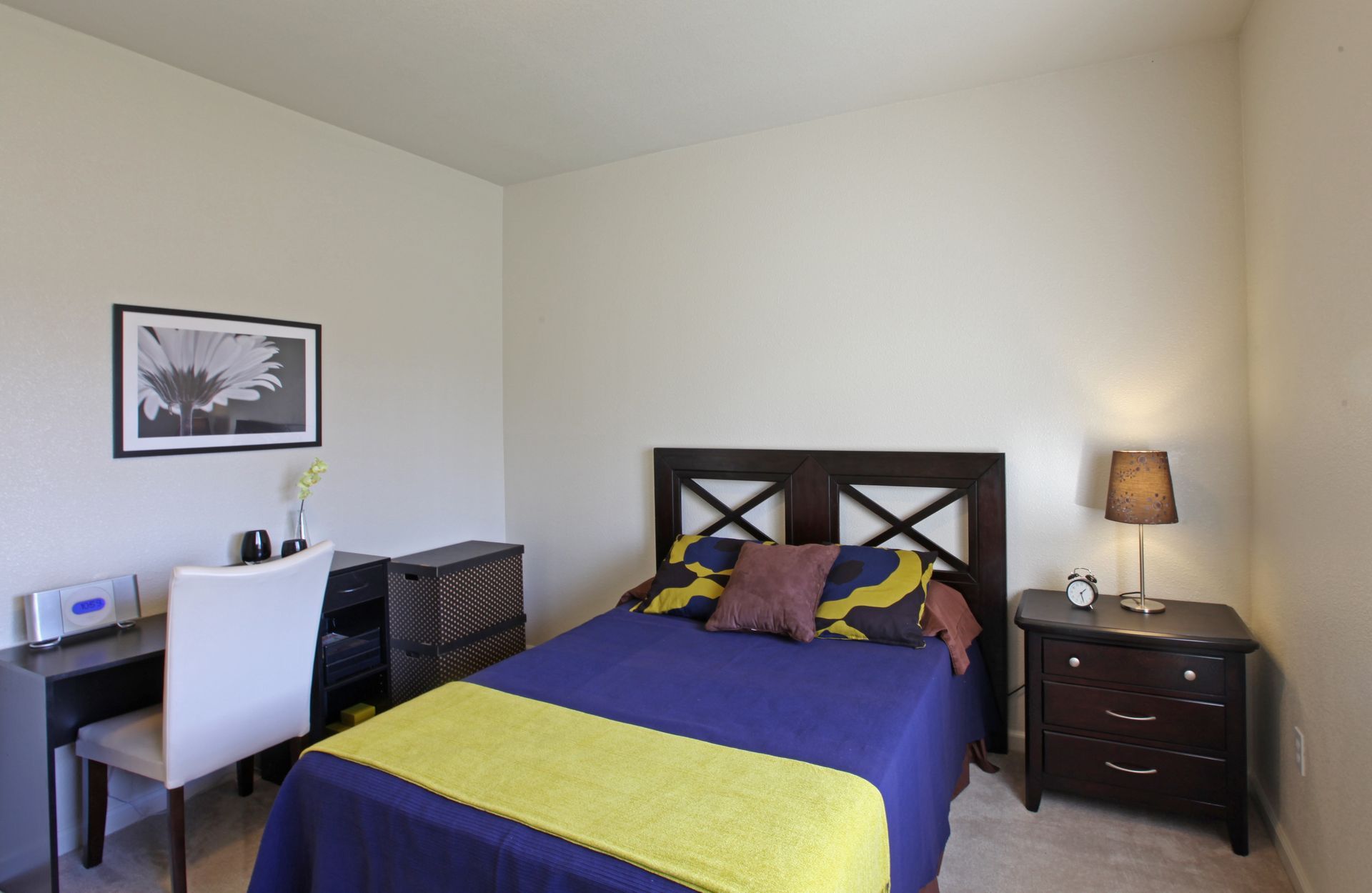 Bedroom with a bed, desk, nightstand, and artwork. Blue and yellow bedding, dark furniture, neutral walls.