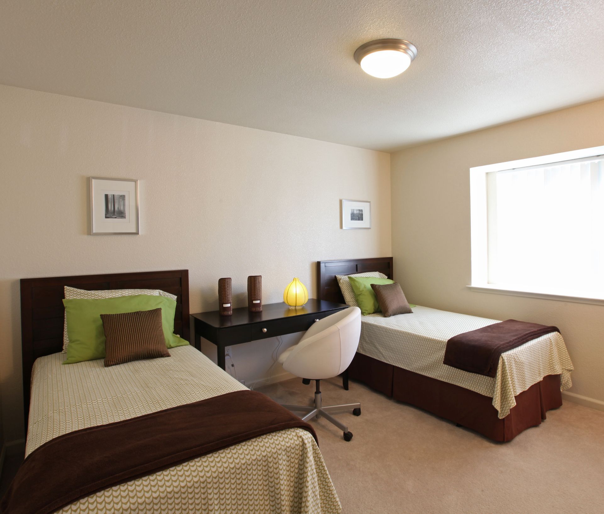 Two twin beds with brown bedding in a beige bedroom; a desk, chair, and artwork are present.