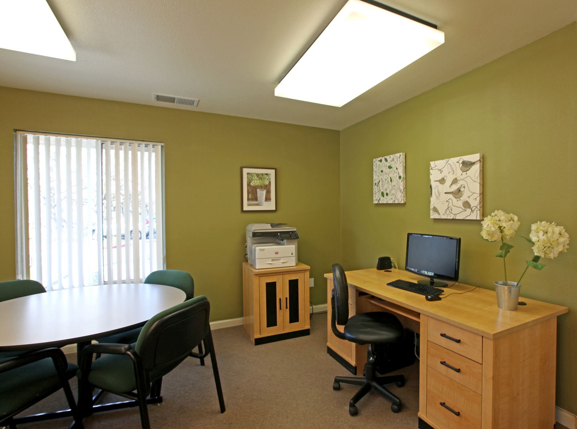 Office with desk, chairs, printer, and a round table; olive green walls and a window with blinds.