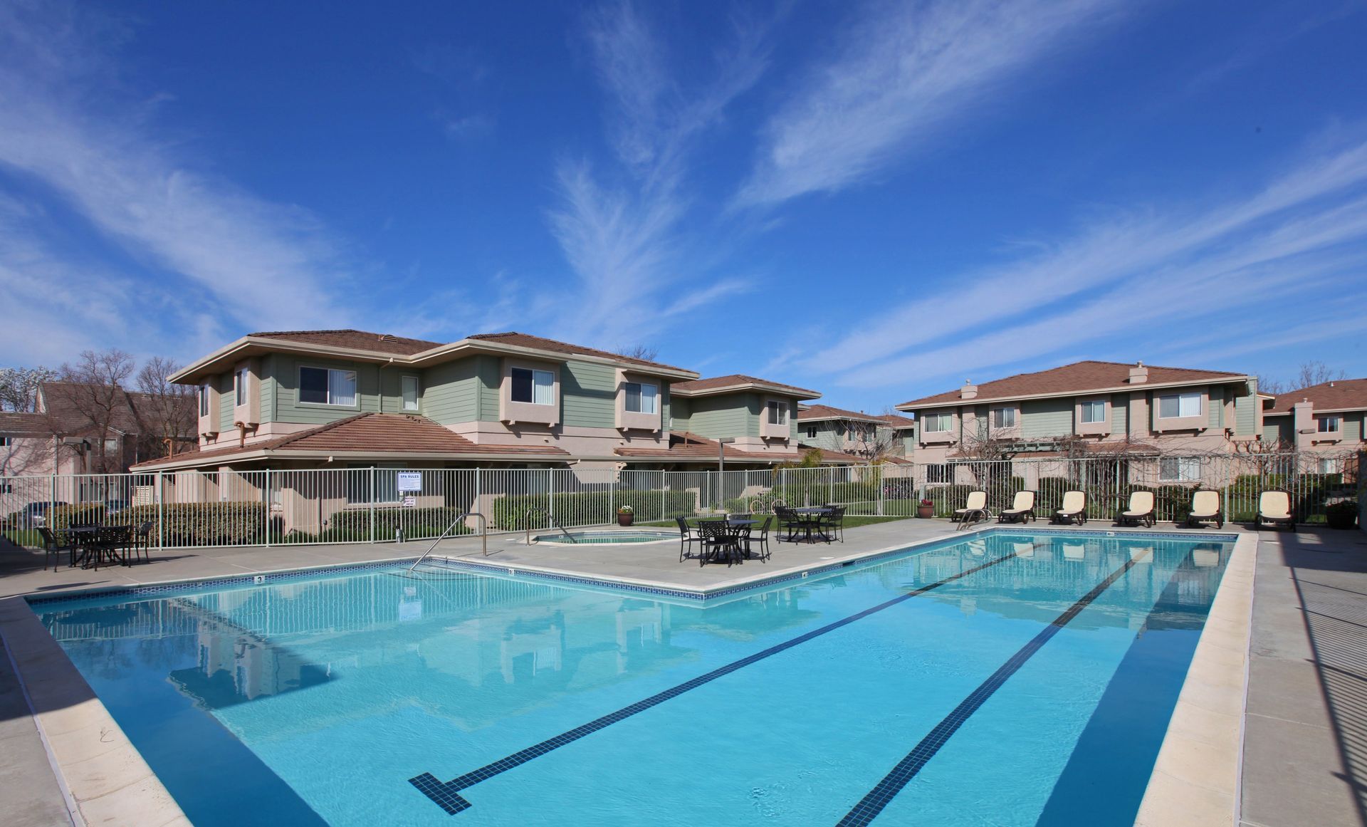 Pool in front of light green apartments with brown roofs under a bright blue sky.