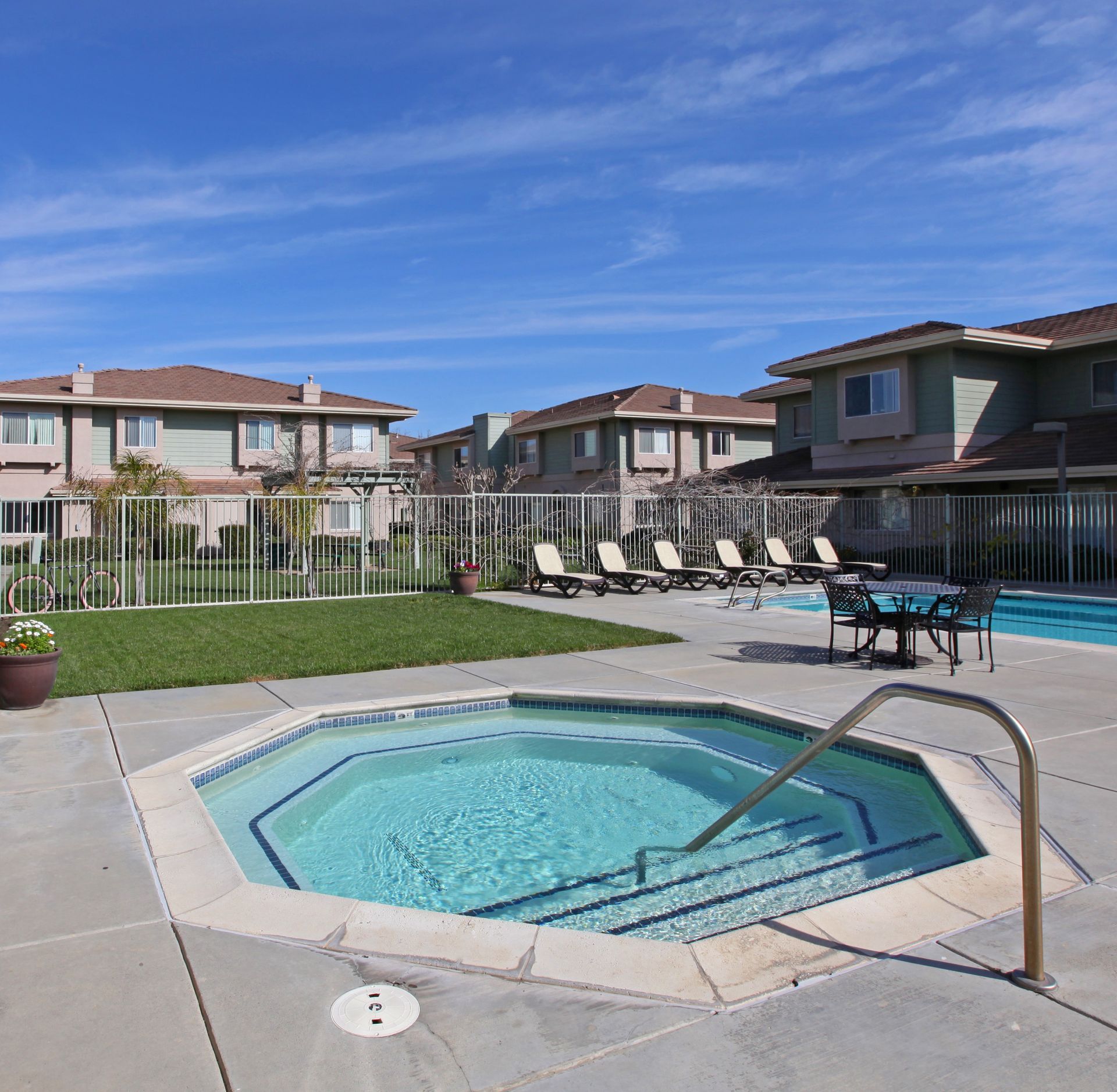 A jacuzzi in front of an apartment building with a swimming pool and lounge chairs on a sunny day.