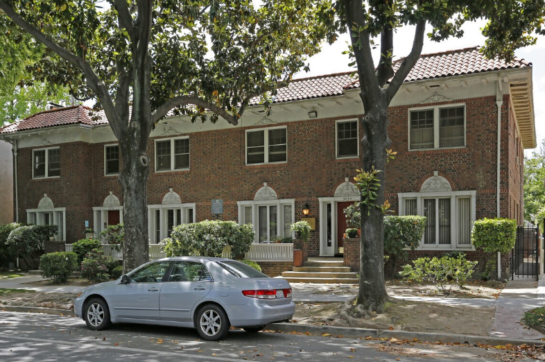 Two-story brick apartment building with a blue sedan parked in front, framed by trees.